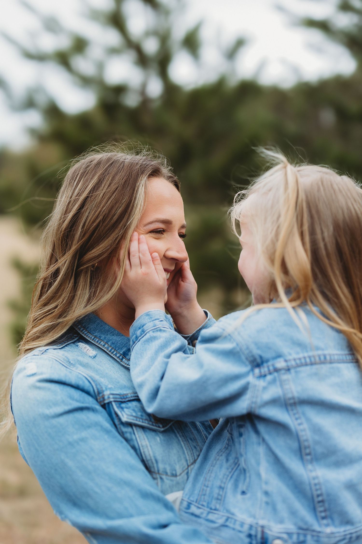 A mother and young daughter in matching denim jackets share a tender moment outdoors, the child gently touching her mom's face.