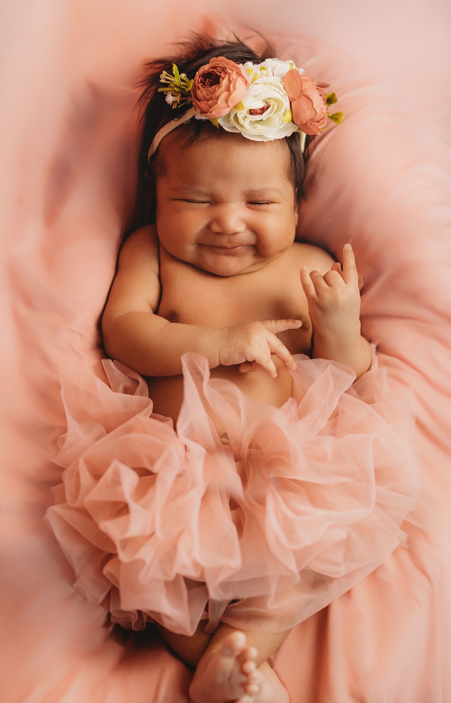 Smiling newborn baby girl wearing a pink tutu and floral headband lying on a soft pink blanket.
