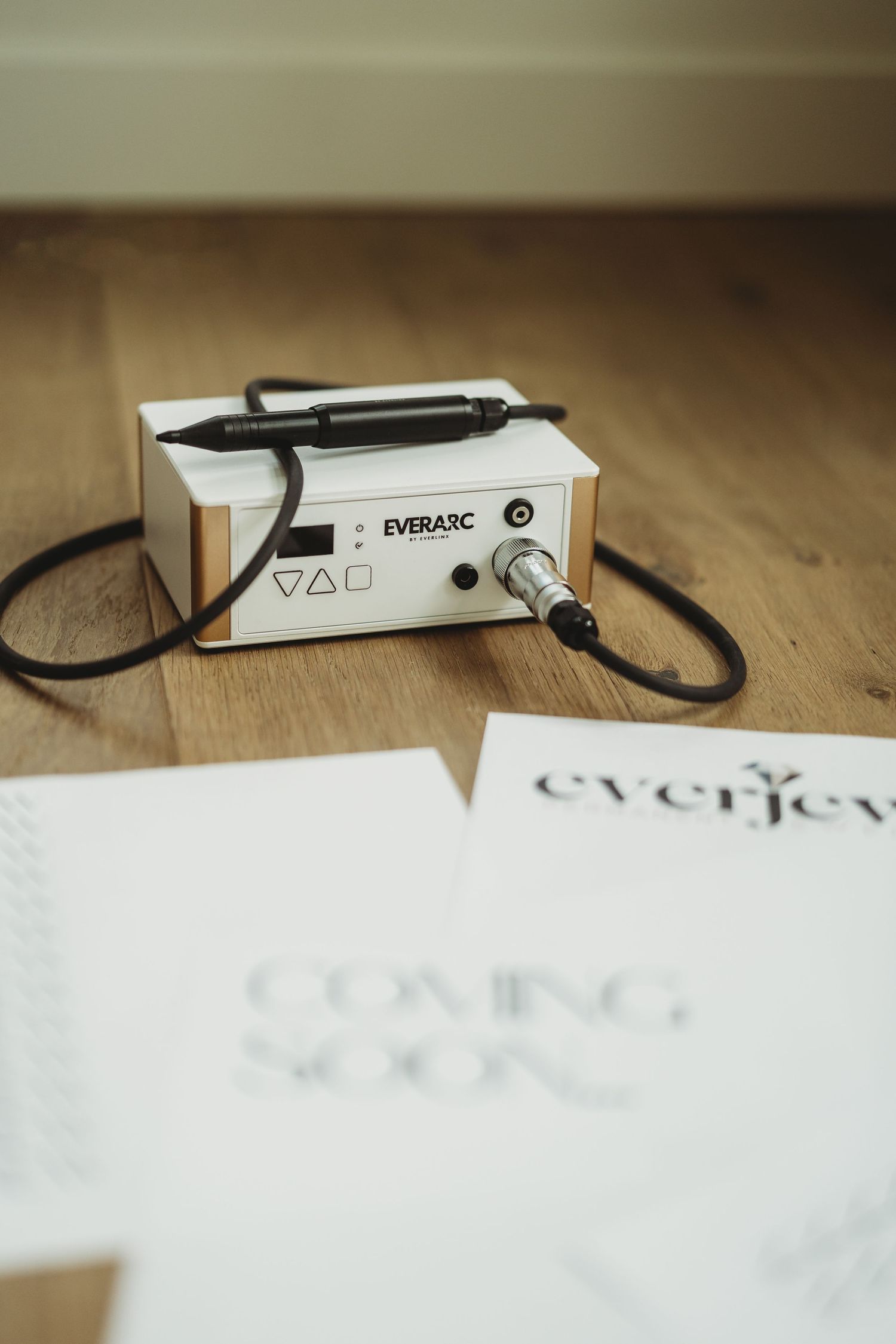 EverArc professional nail drill machine with handpiece on wooden floor next to Coming Soon branding papers.