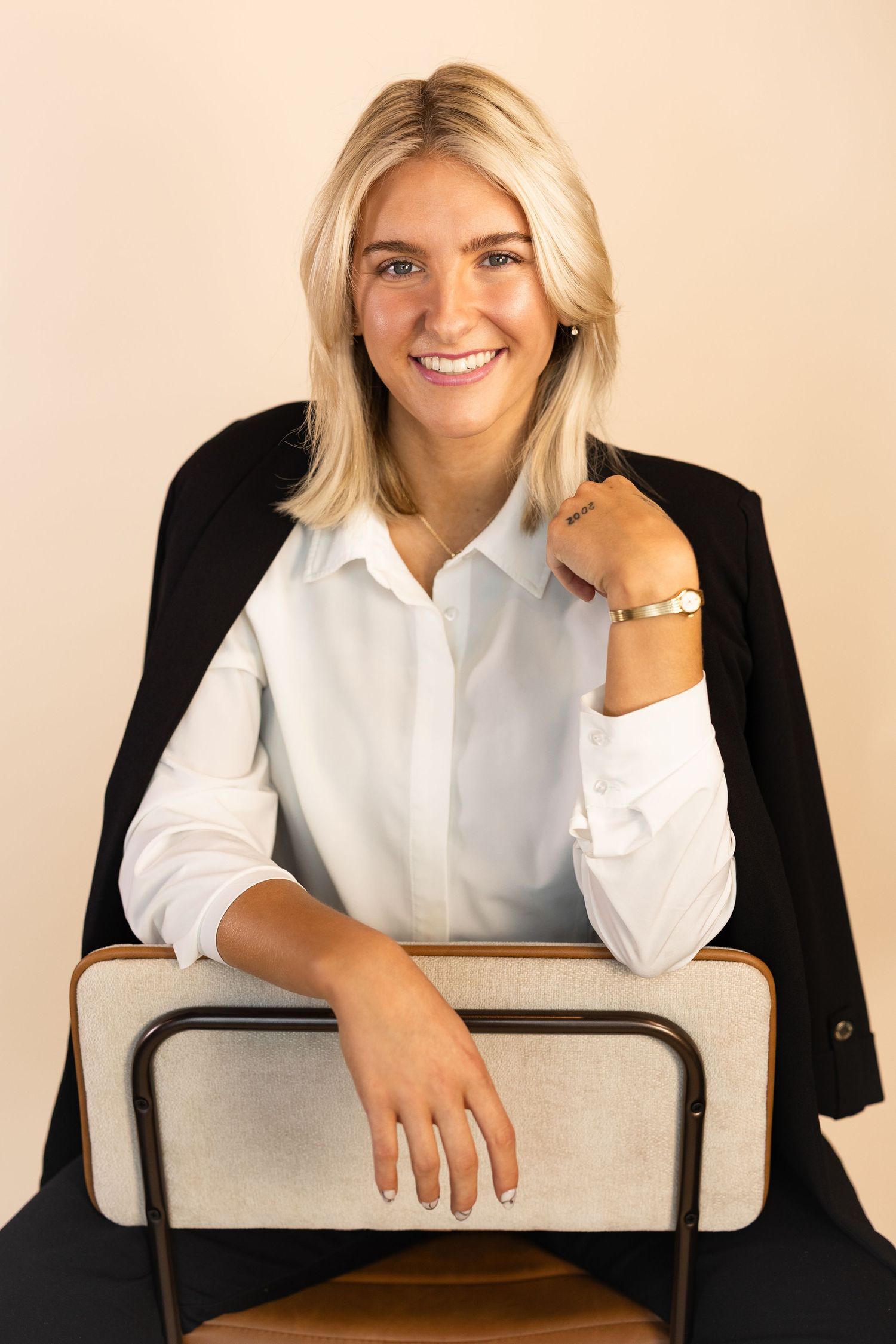 Smiling blonde woman in white shirt and black blazer posing on chair with gold bracelet accessory.
