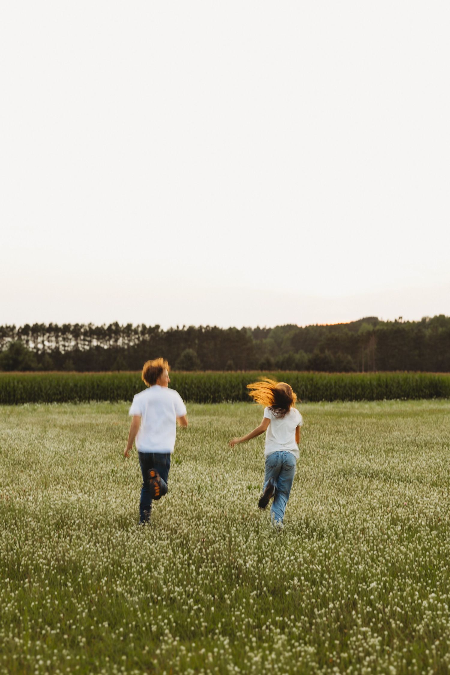 Two children in white shirts and jeans running through a blooming meadow with trees in the background at dusk.