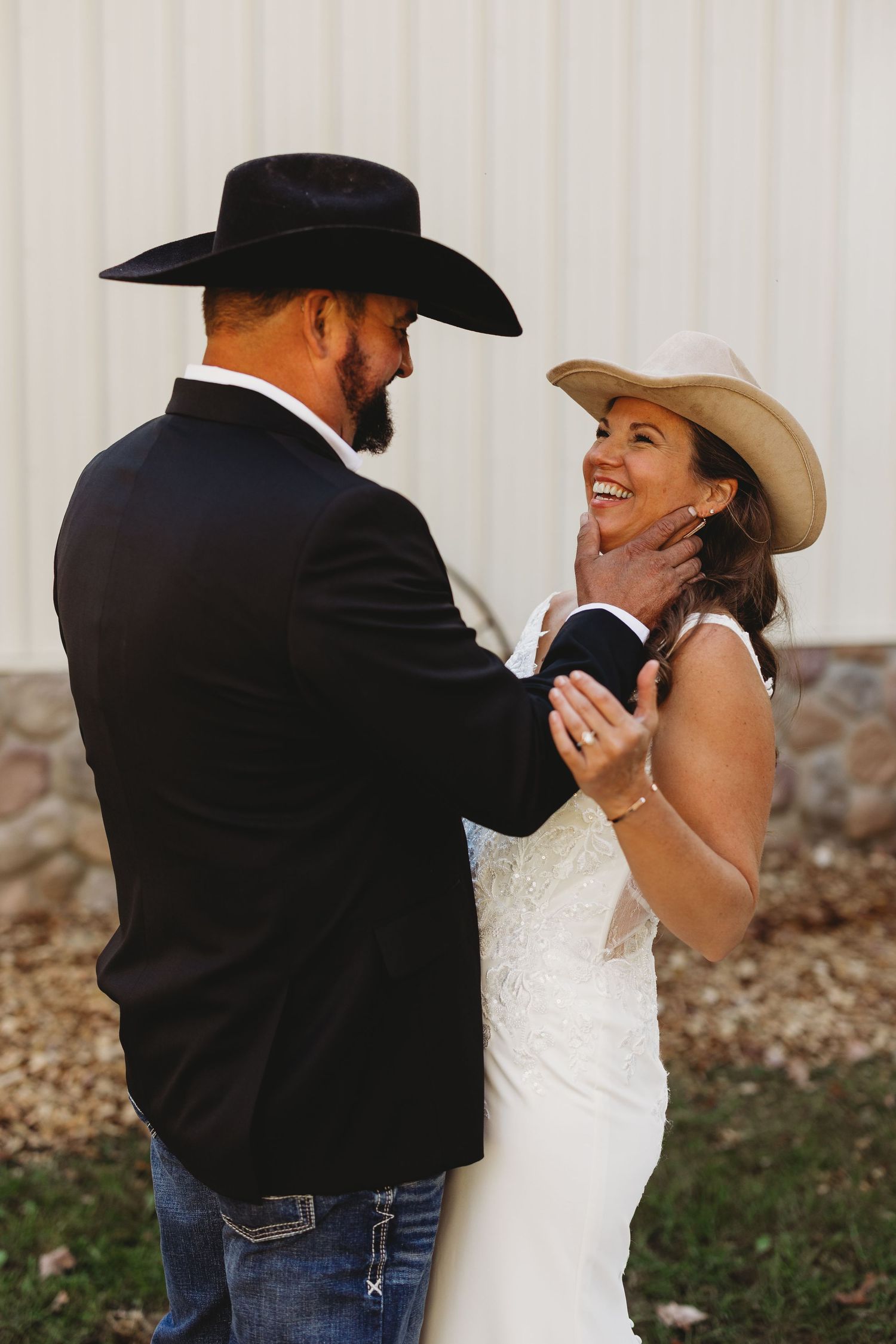 Smiling bride in a lace wedding dress and tan cowboy hat laughs as her groom in a black hat gently touches her face.