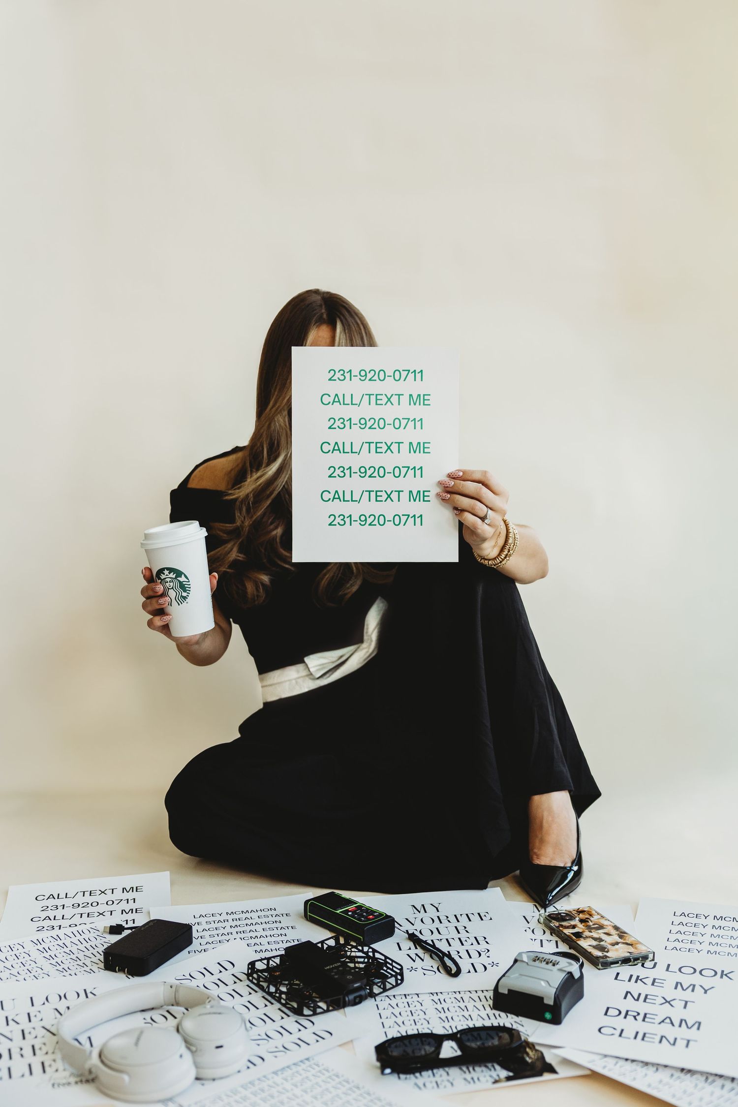 Woman in black outfit sits on the floor holding a Starbucks cup and a call or text me sign over her face, surrounded by phones and papers.