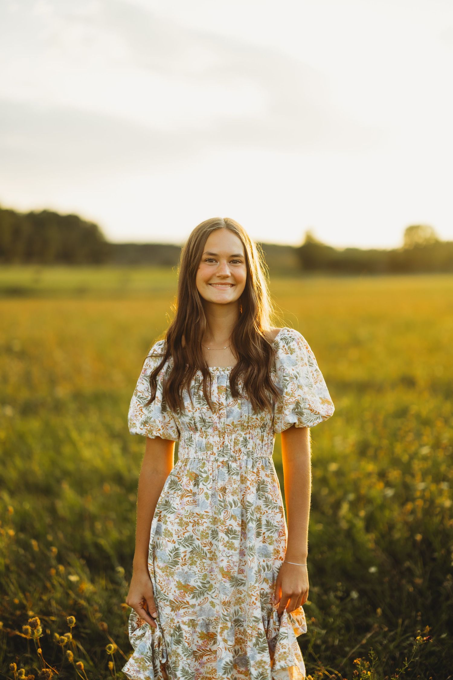 Smiling young woman in a floral puff-sleeve dress stands in a golden wildflower meadow at sunset.