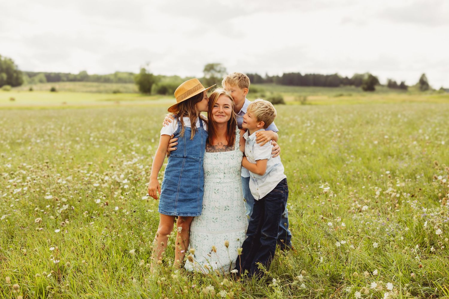 A smiling mother hugs three children in a sunny wildflower meadow, with the kids in a straw hat and casual clothes.