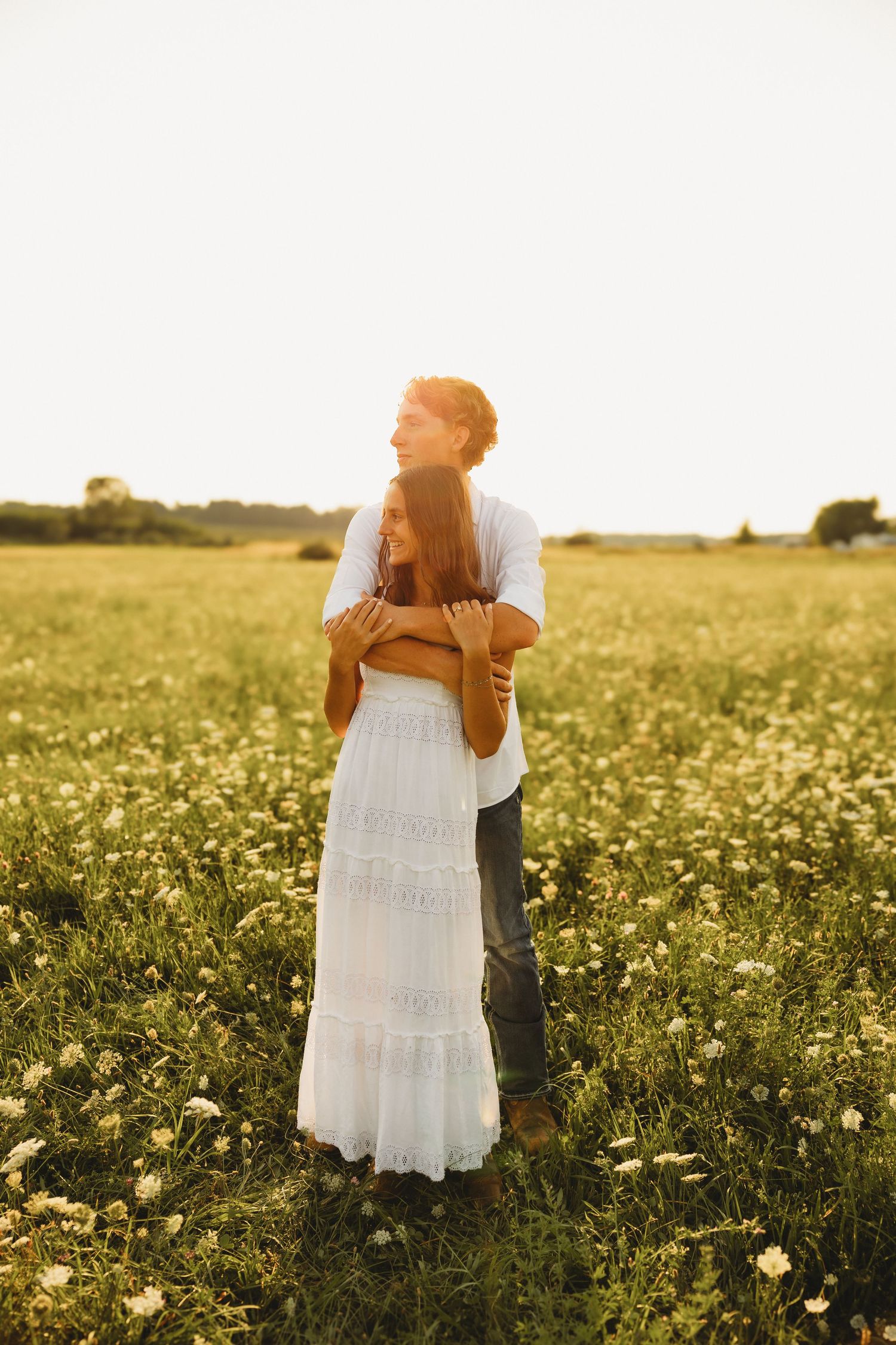 Couple embracing in a golden sunlit wildflower field, woman in white boho dress, romantic outdoor portrait.