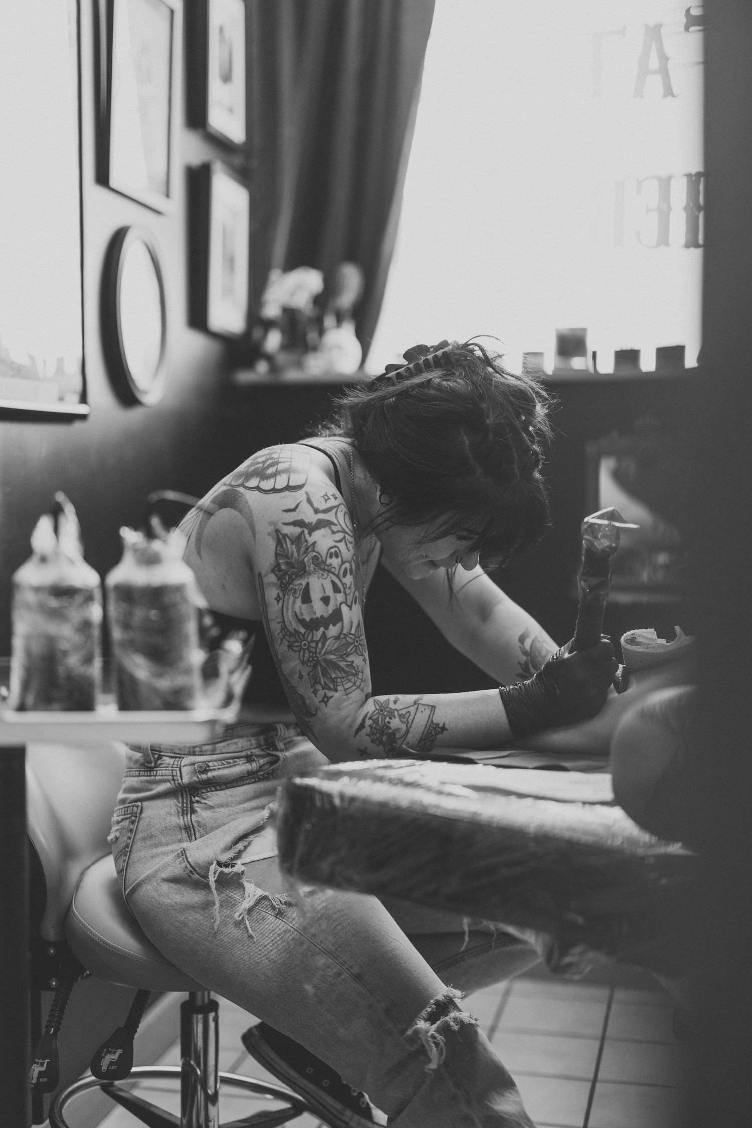 Black and white photo of a tattooed female tattoo artist working on a client in a tattoo parlor.