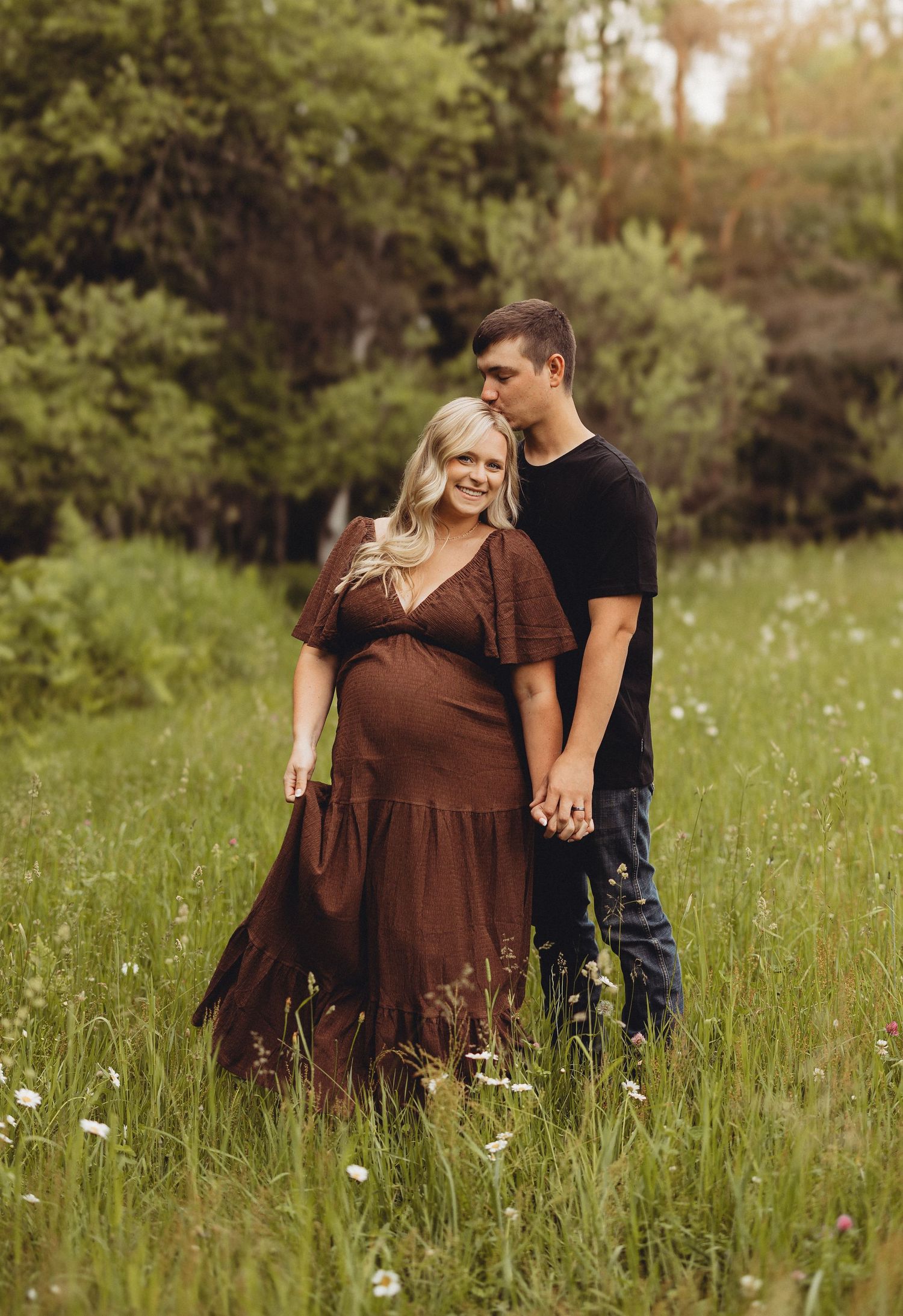 Pregnant couple holding hands in a lush green meadow, woman in brown maternity dress, man in black shirt and jeans.