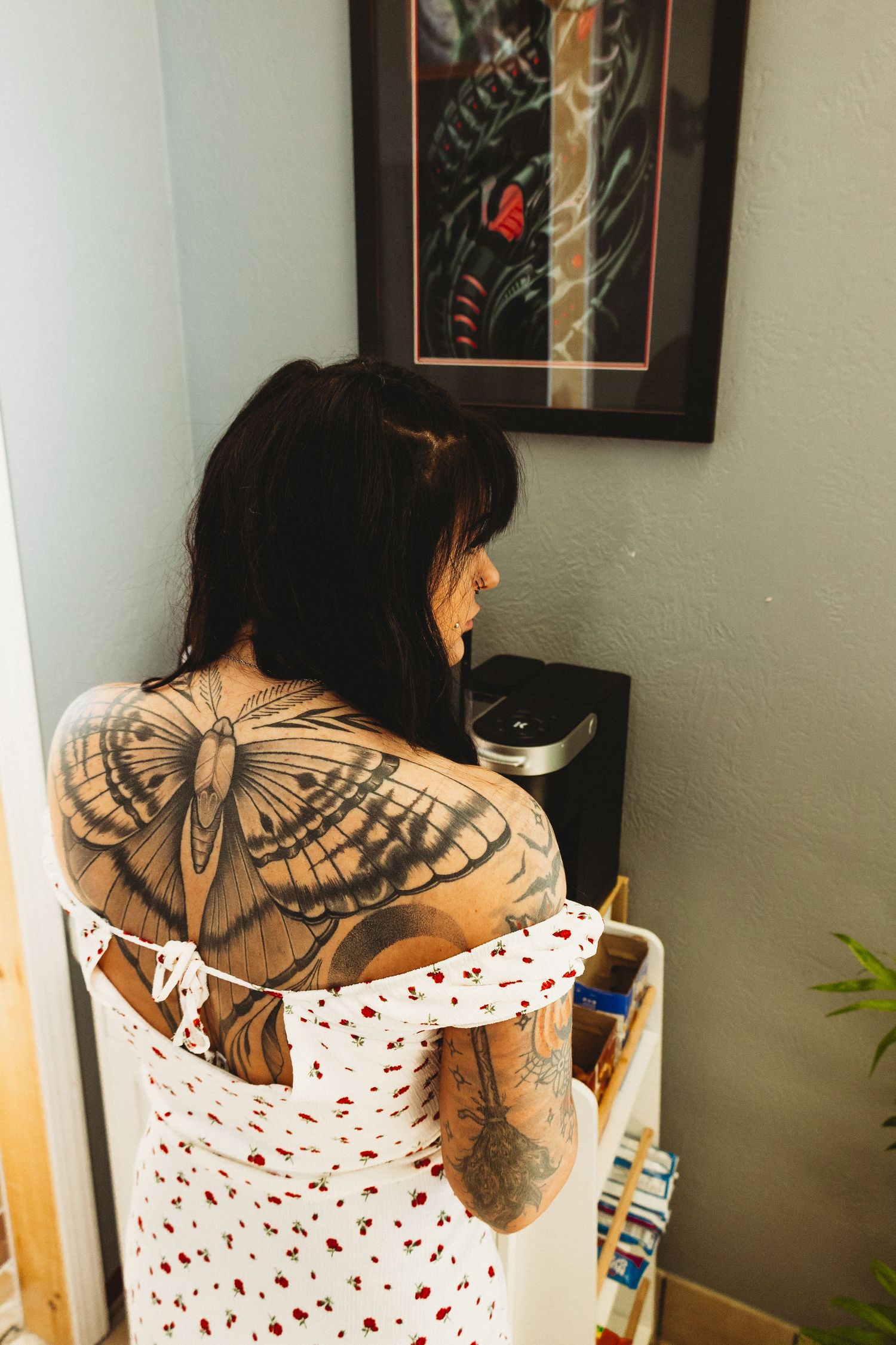 Woman with large butterfly back tattoo viewed from behind near a framed tattoo print.