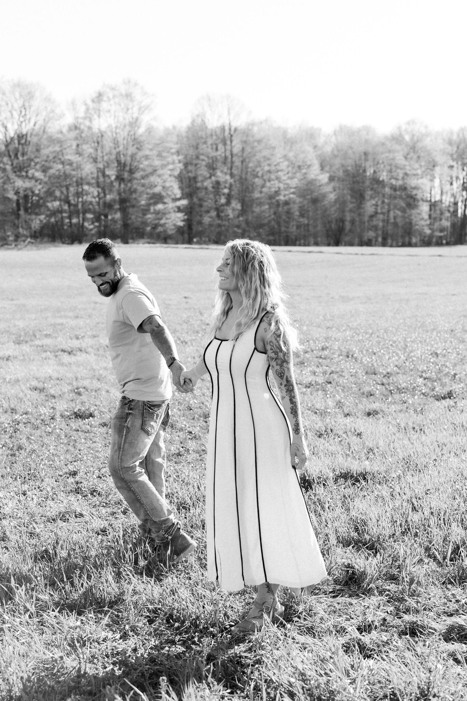 Black and white photo of a tattooed couple holding hands while walking through an open field with trees in the background.