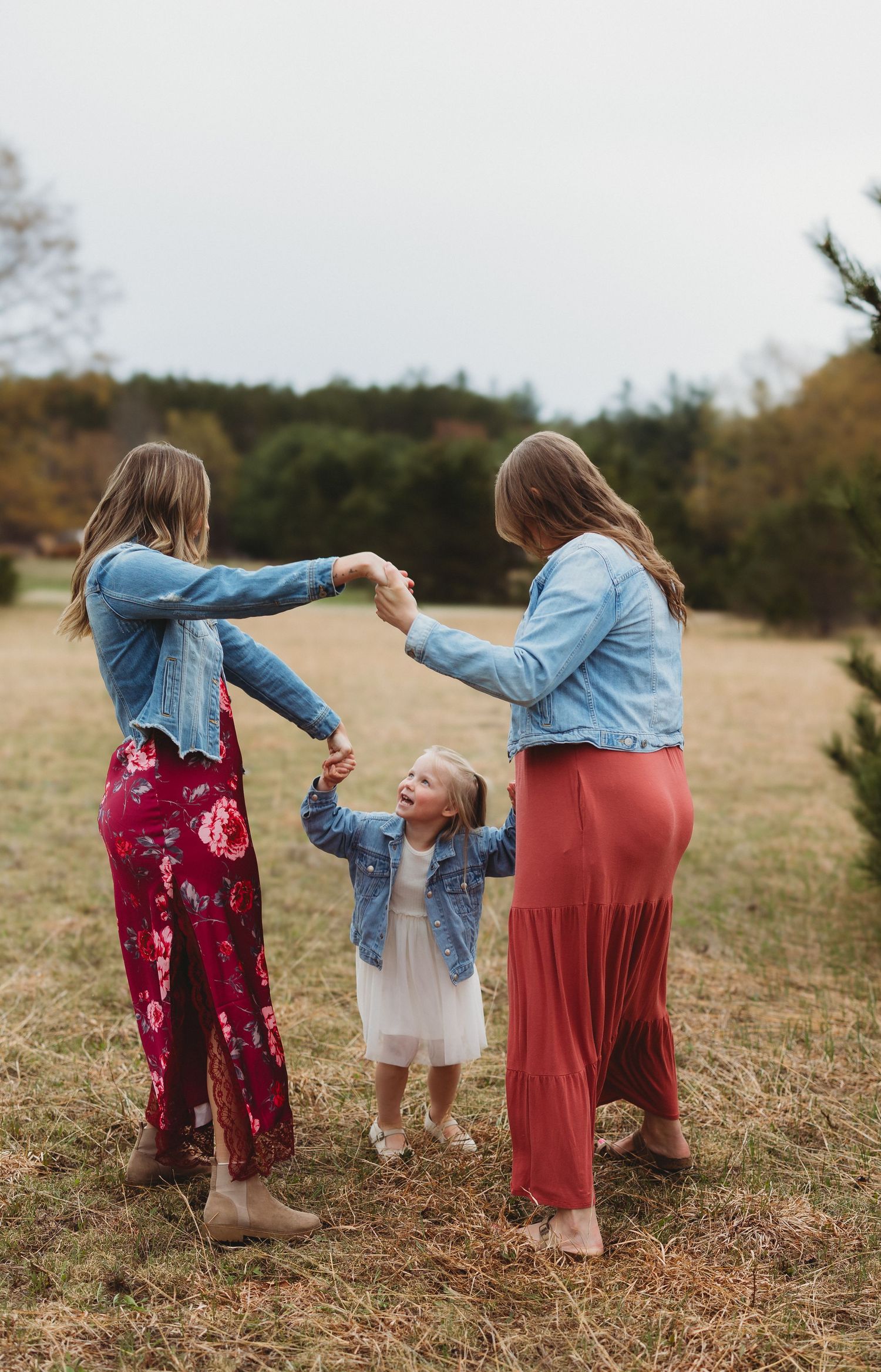 Two women in denim jackets and maxi skirts hold hands with a young girl in a white dress in an outdoor field.