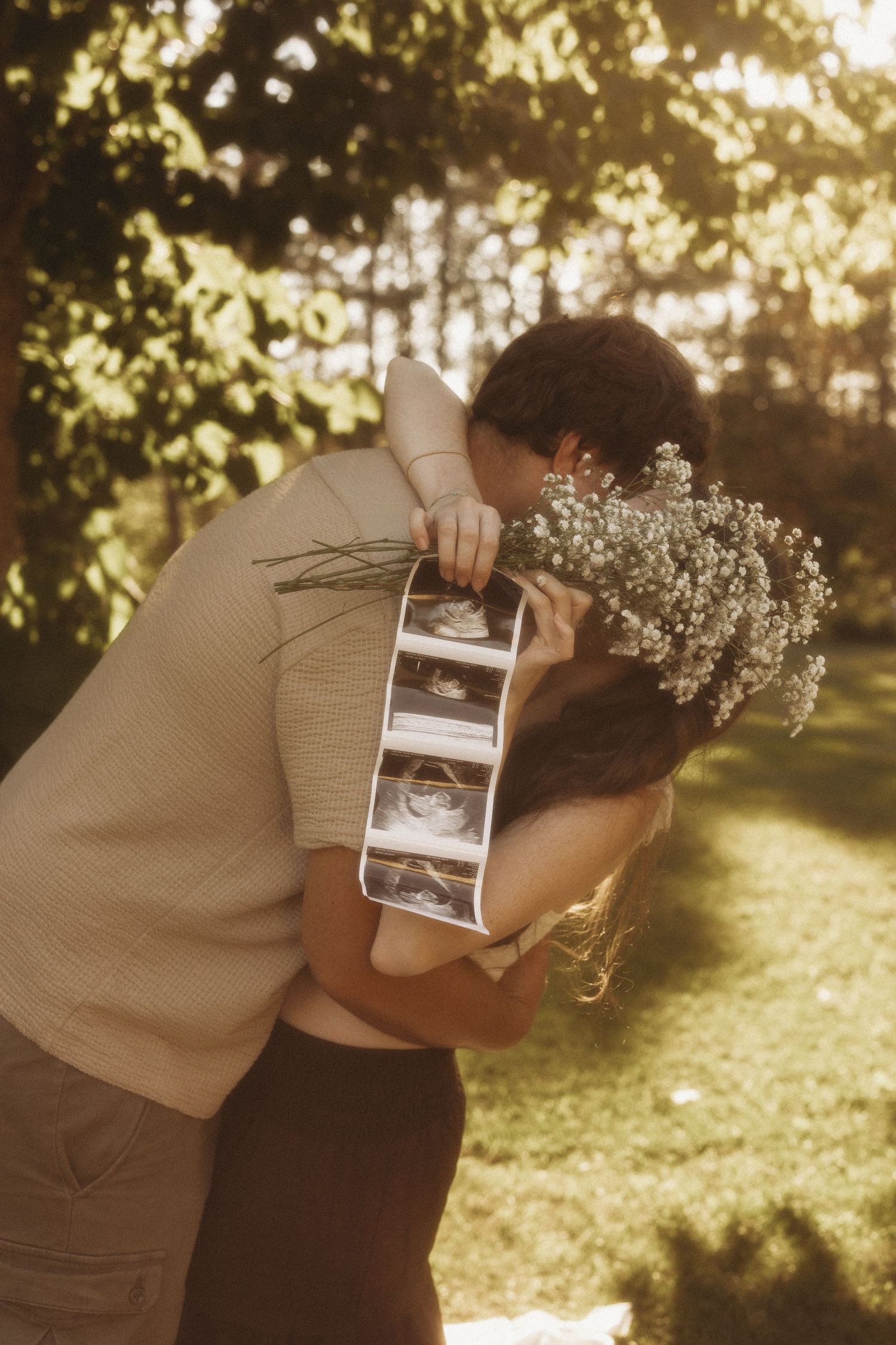 Couple embracing outdoors holding ultrasound photos and wildflowers in golden sunlight, announcing pregnancy in a garden setting.