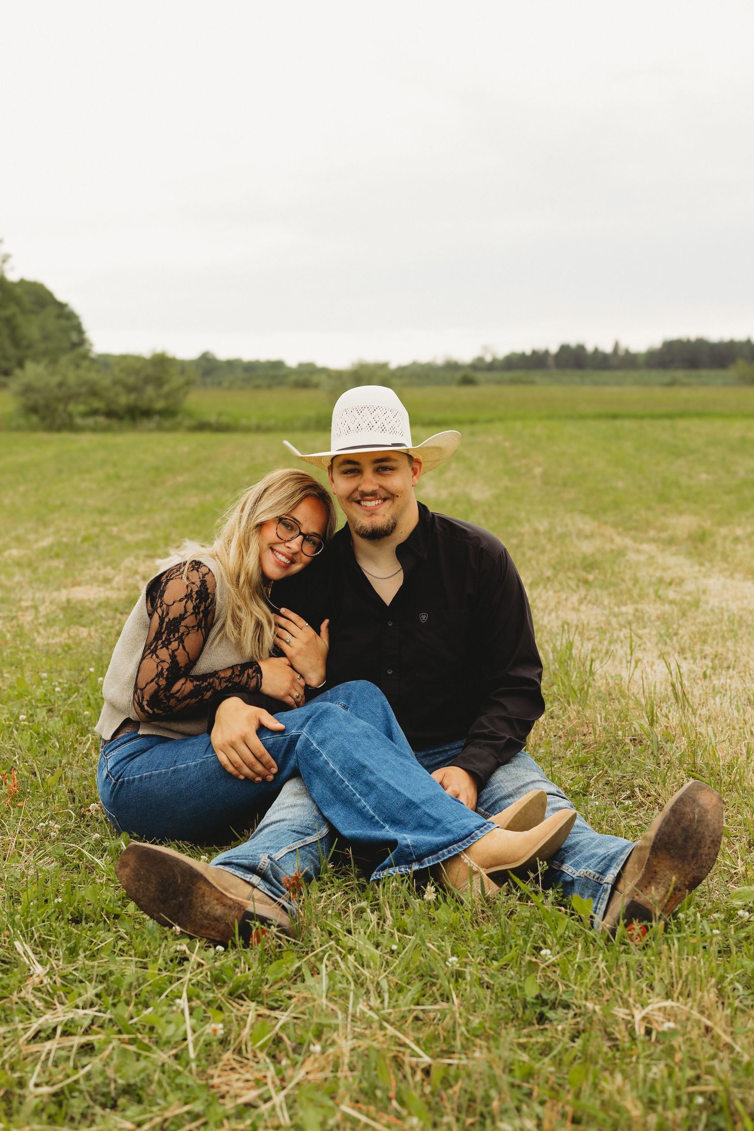 A smiling couple sits together on green grass in a field, the man wearing a cowboy hat and jeans, the woman leaning on his shoulder.
