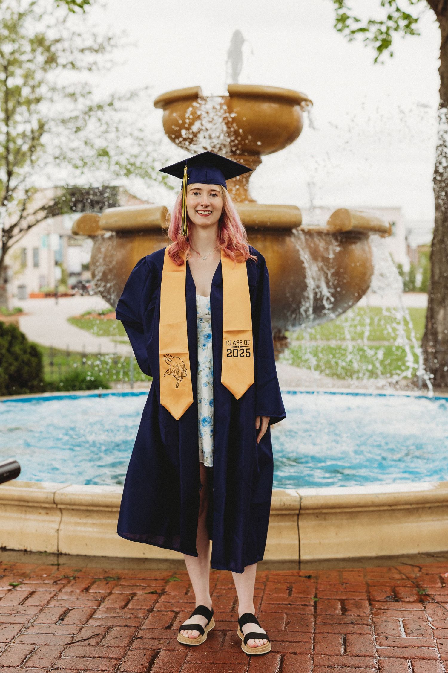 Graduate in academic regalia poses by fountain wearing navy cap and gold stole.