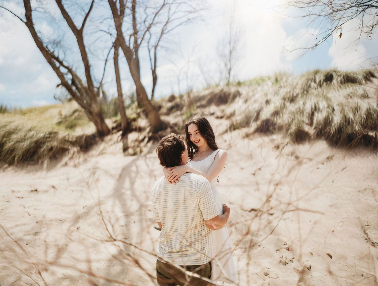 Couple embracing on a sandy beach with driftwood and dunes in the background.