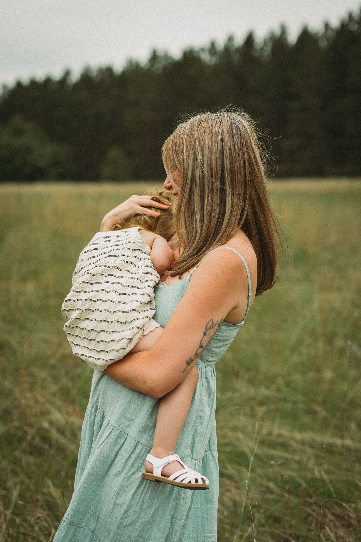 A tender embrace between parent and child in a mint green dress standing in a serene meadow with evergreen trees.