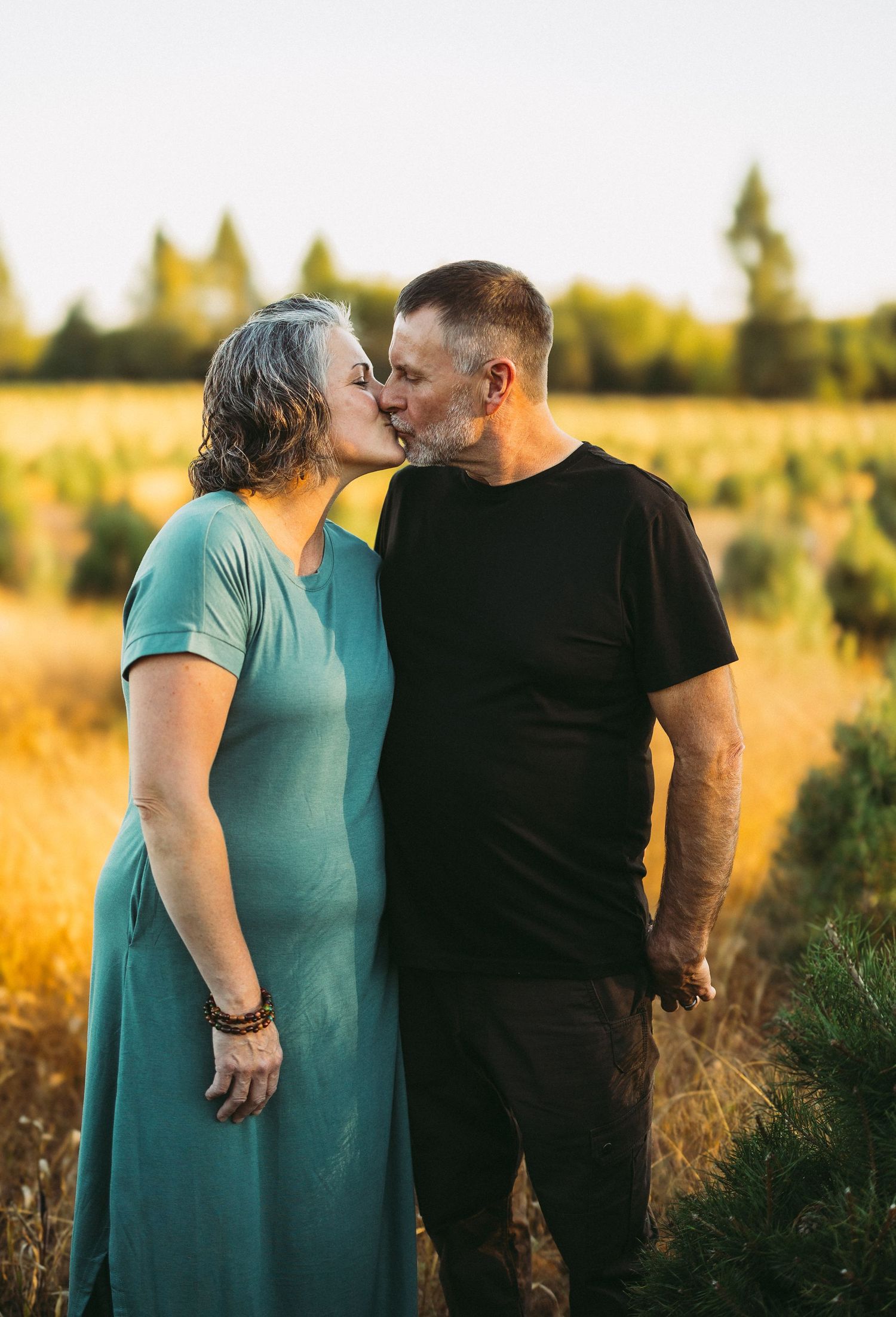 A couple shares a tender moment in a sunlit field during golden hour, wearing teal and black outfits.