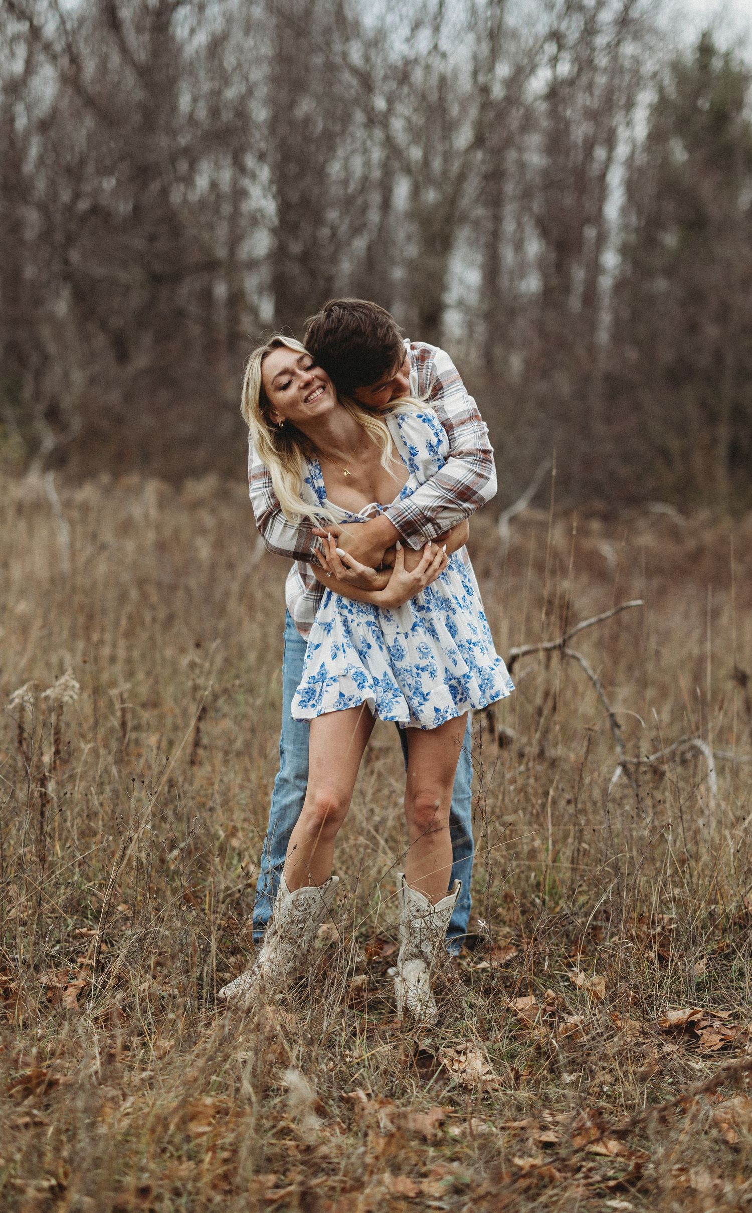 A couple embraces in a rustic field with tall grass and bare trees on an autumn day.
