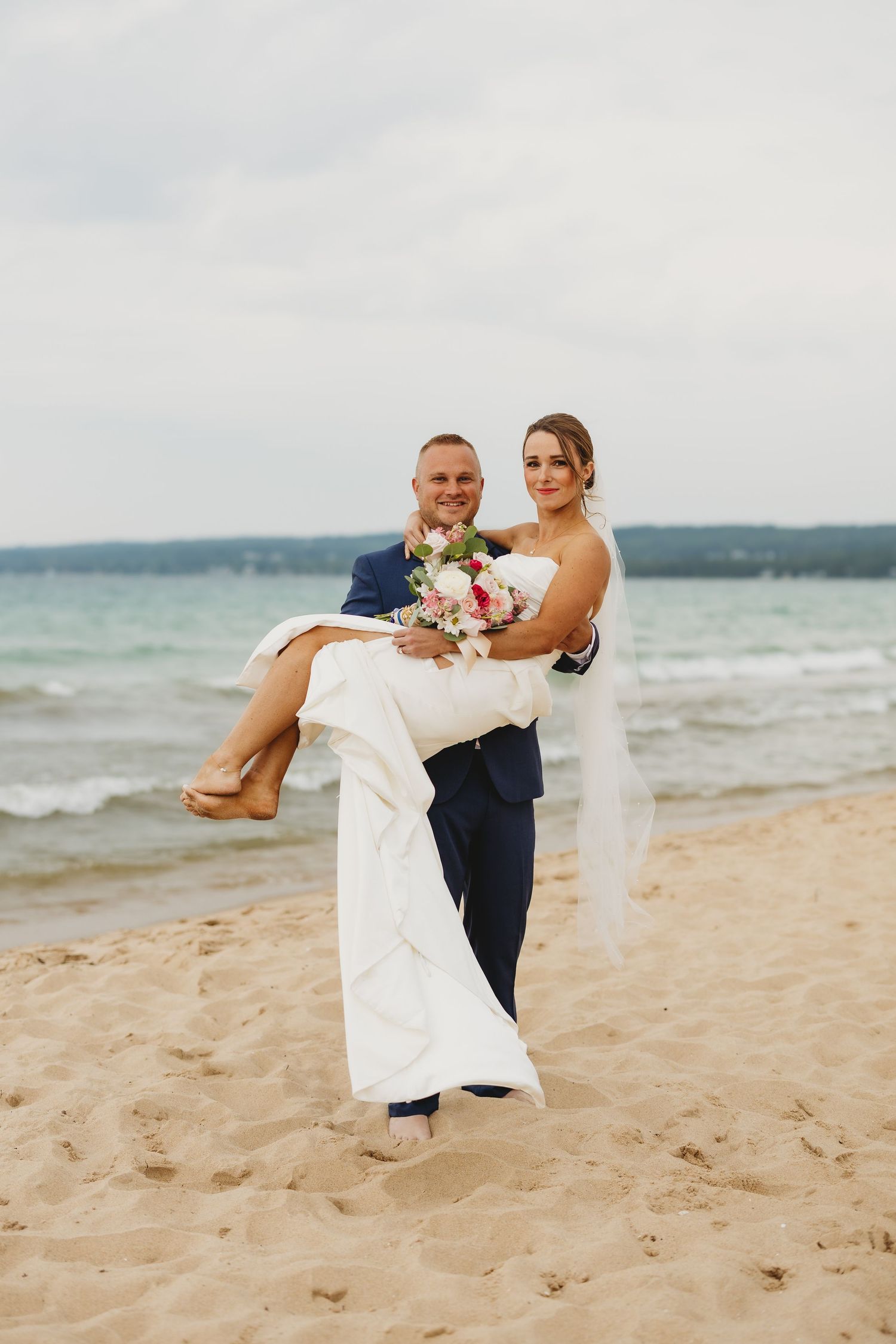 Groom carries bride in white dress and bouquet on sandy beach with ocean backdrop on their wedding day.