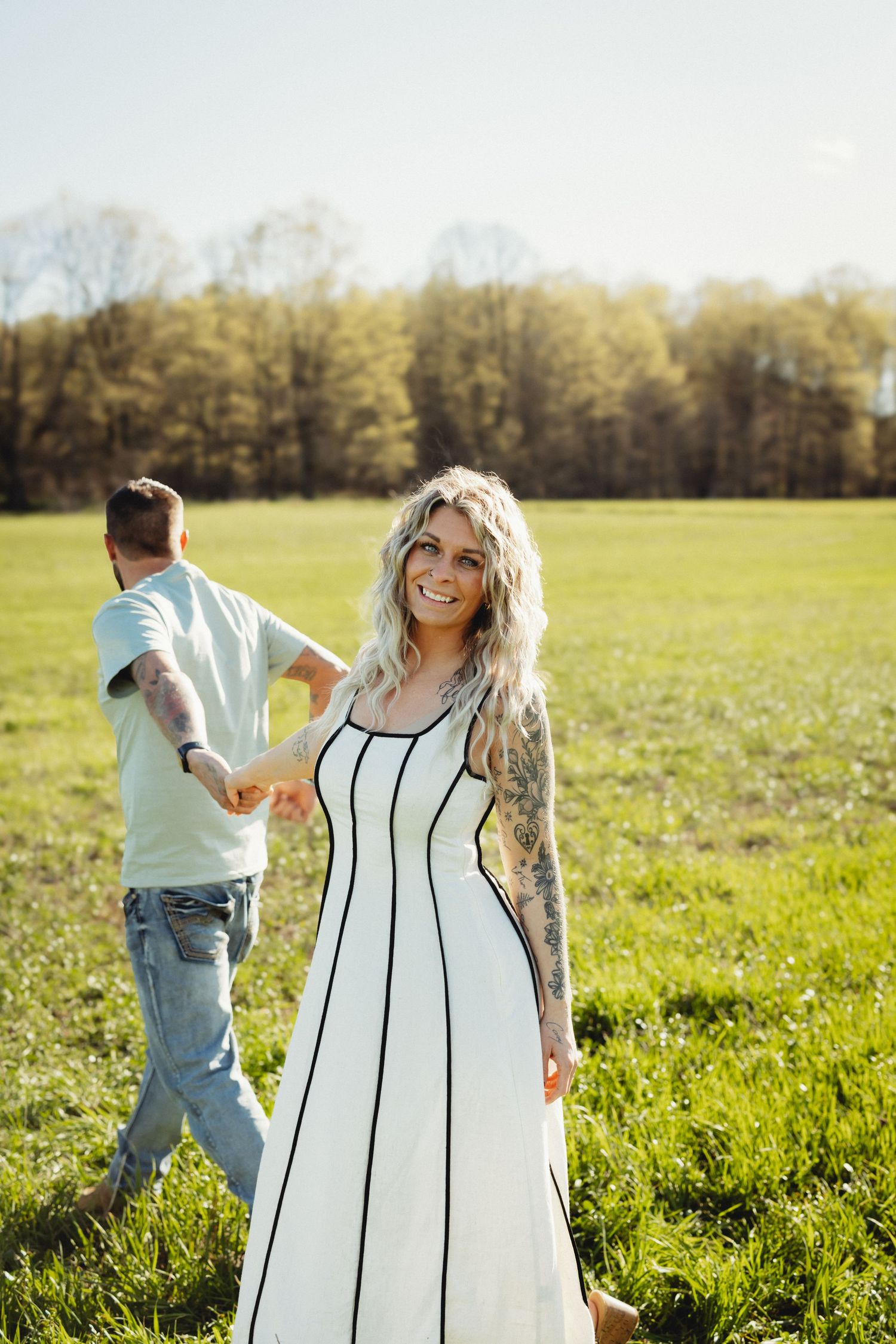 Tattooed blonde woman in white dress holds hands with man in green field on sunny day.