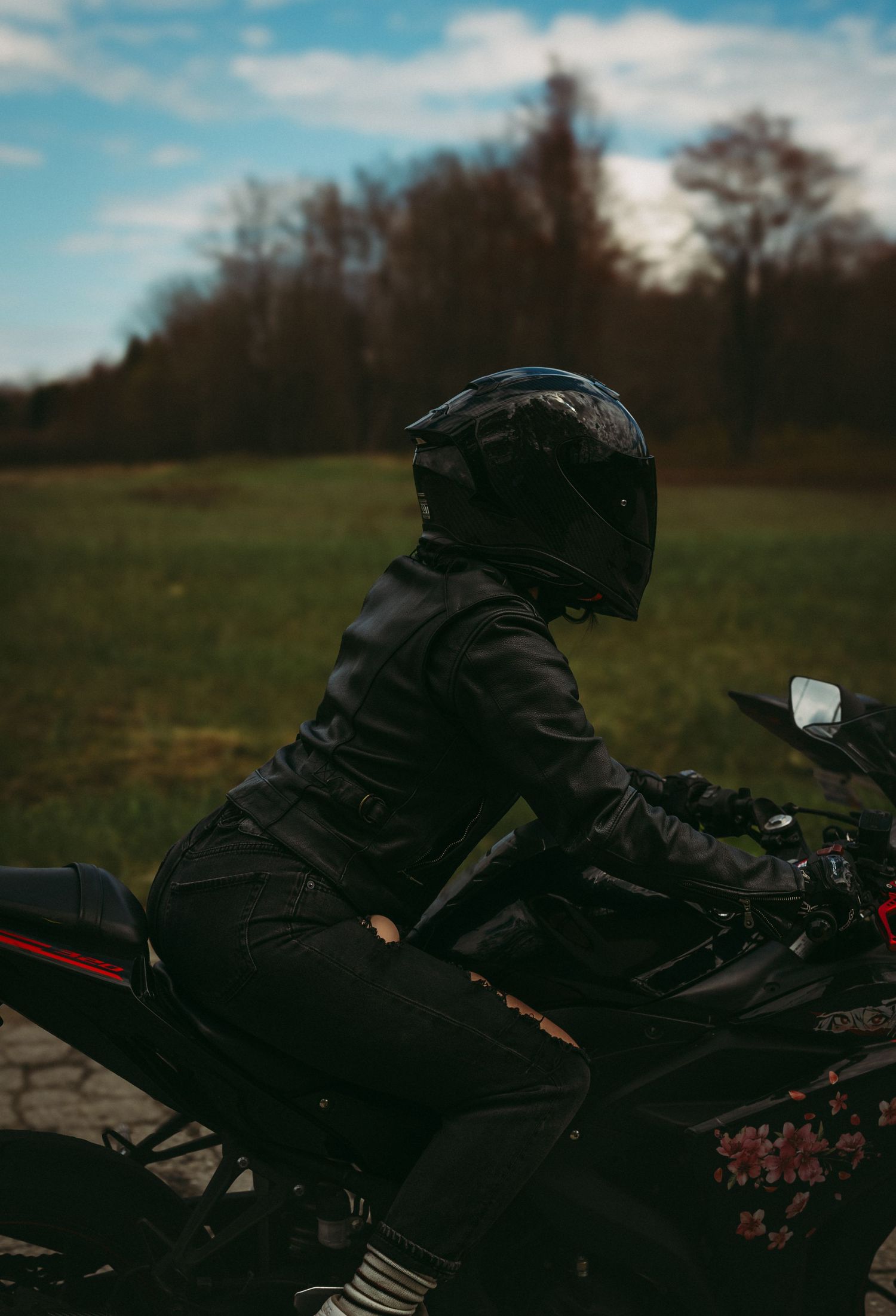 Motorcyclist in black helmet and jacket sitting on a sport motorcycle in a scenic outdoor field with trees and cloudy sky.