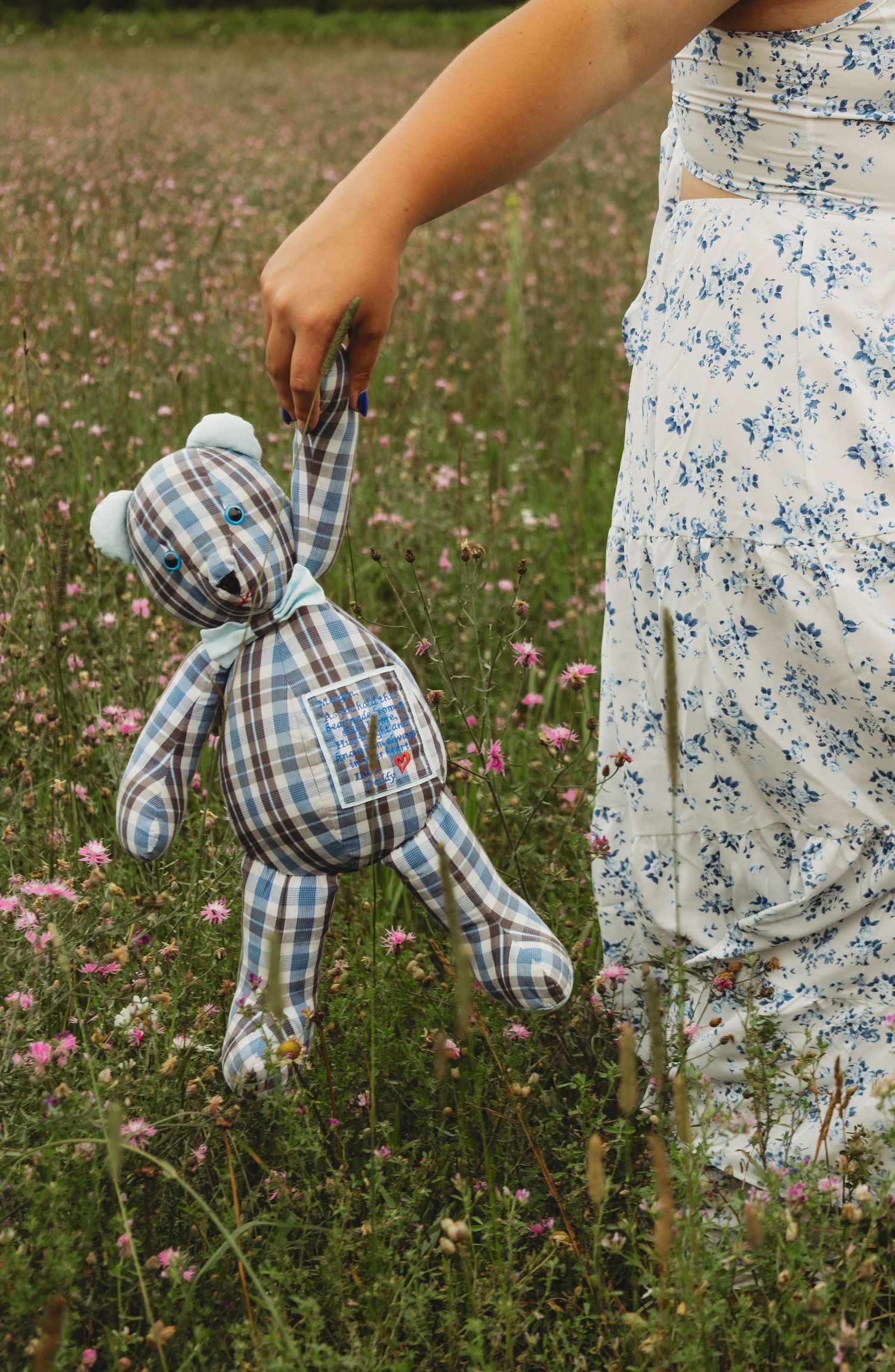 A girl in a floral dress holds a handmade plaid teddy bear in a wildflower meadow with pink blooms.