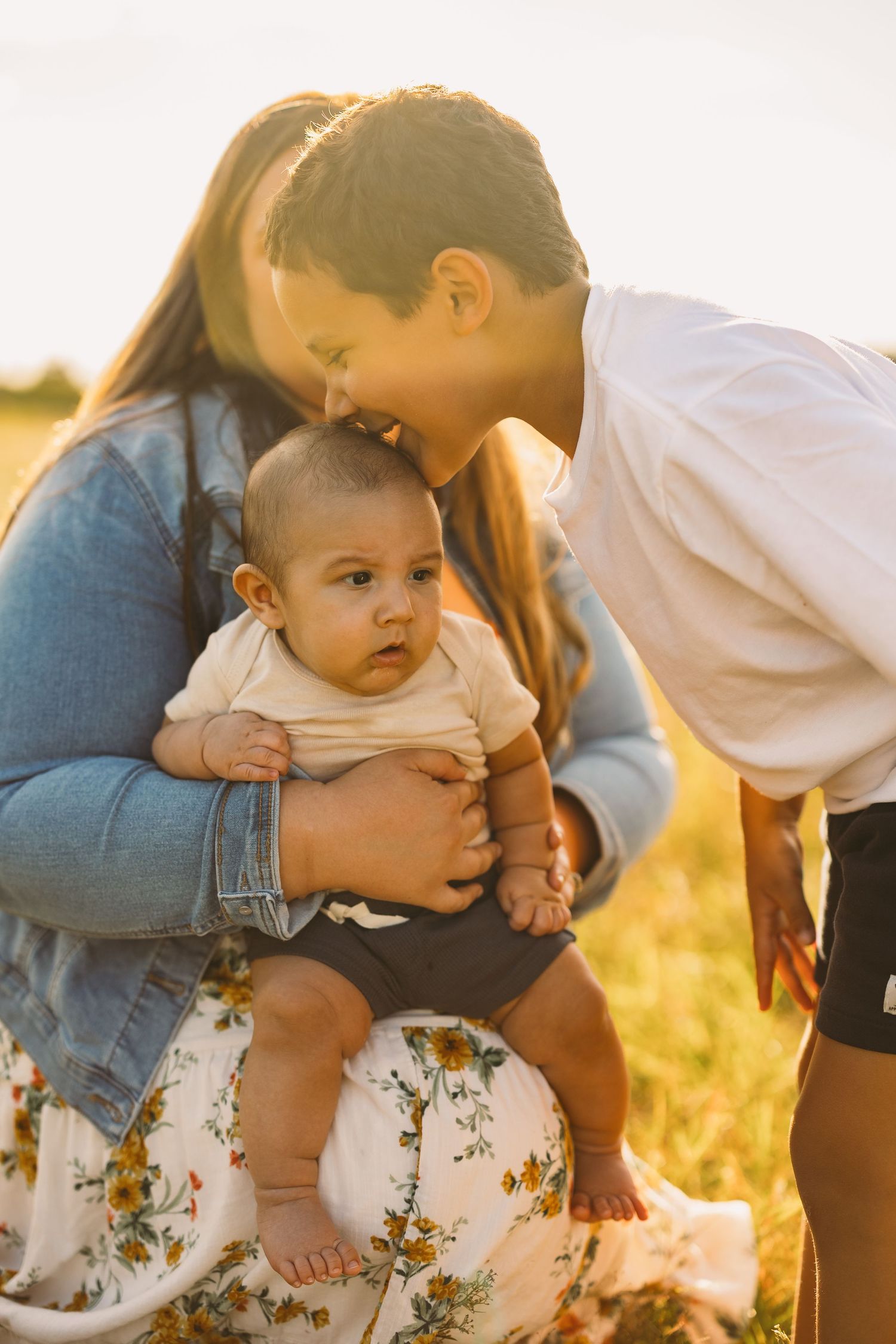 Smiling boy kisses a baby held by a woman in a denim jacket and floral skirt during a golden-hour family photo session in a sunny field.