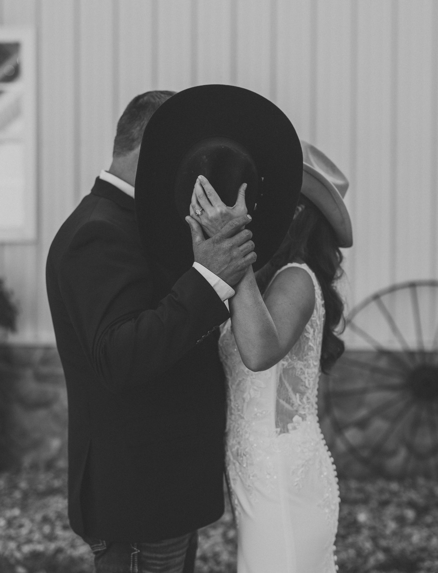 Black and white photo of a groom in a suit and a bride in a lace gown kissing behind a black cowboy hat at a rustic barn wedding.