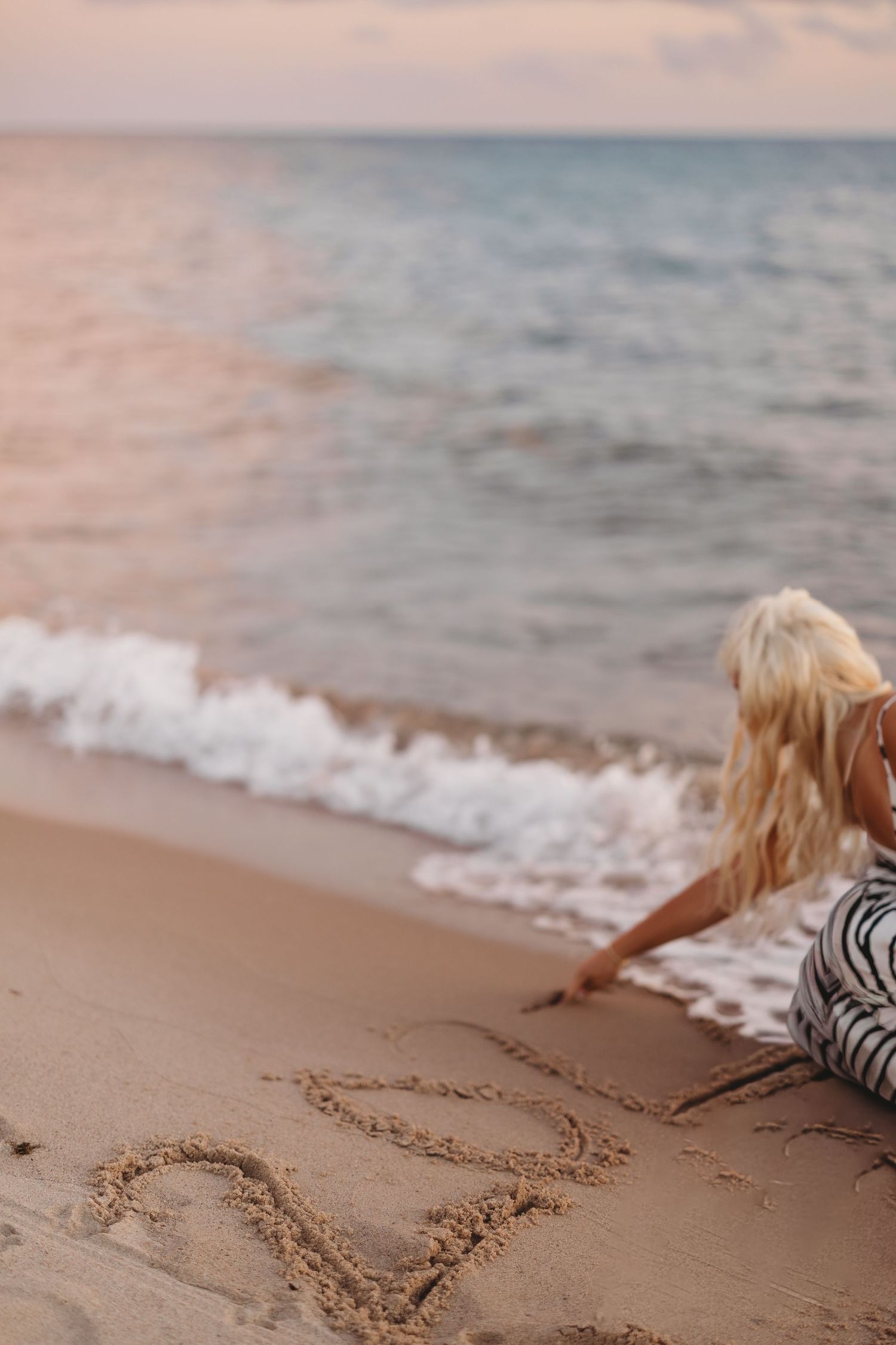 Blonde woman in a striped dress sits on a sandy beach at sunset, writing in the sand beside gentle ocean waves.