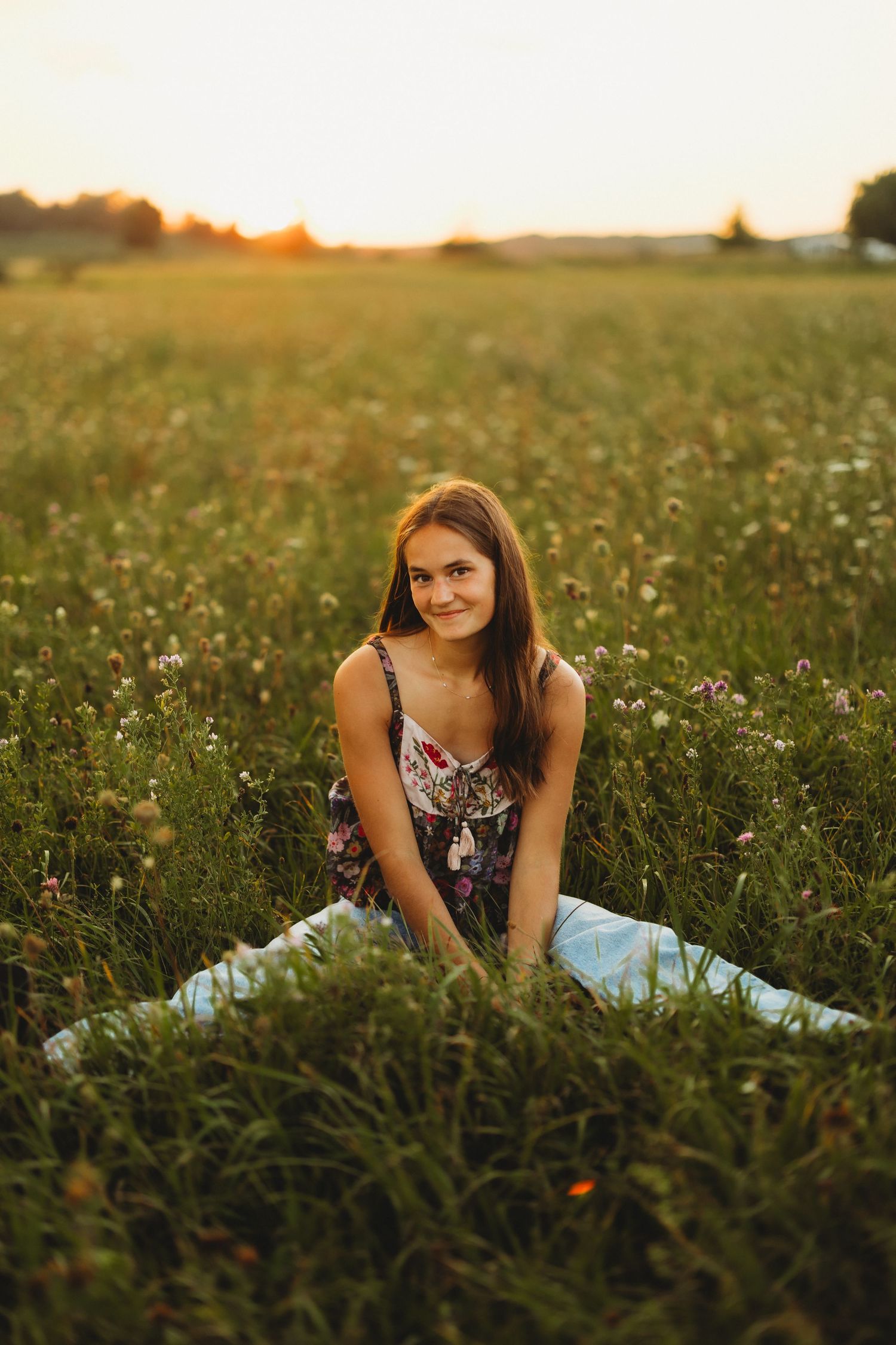 Smiling young woman with long brown hair sits cross-legged in a wildflower meadow at golden hour sunset.