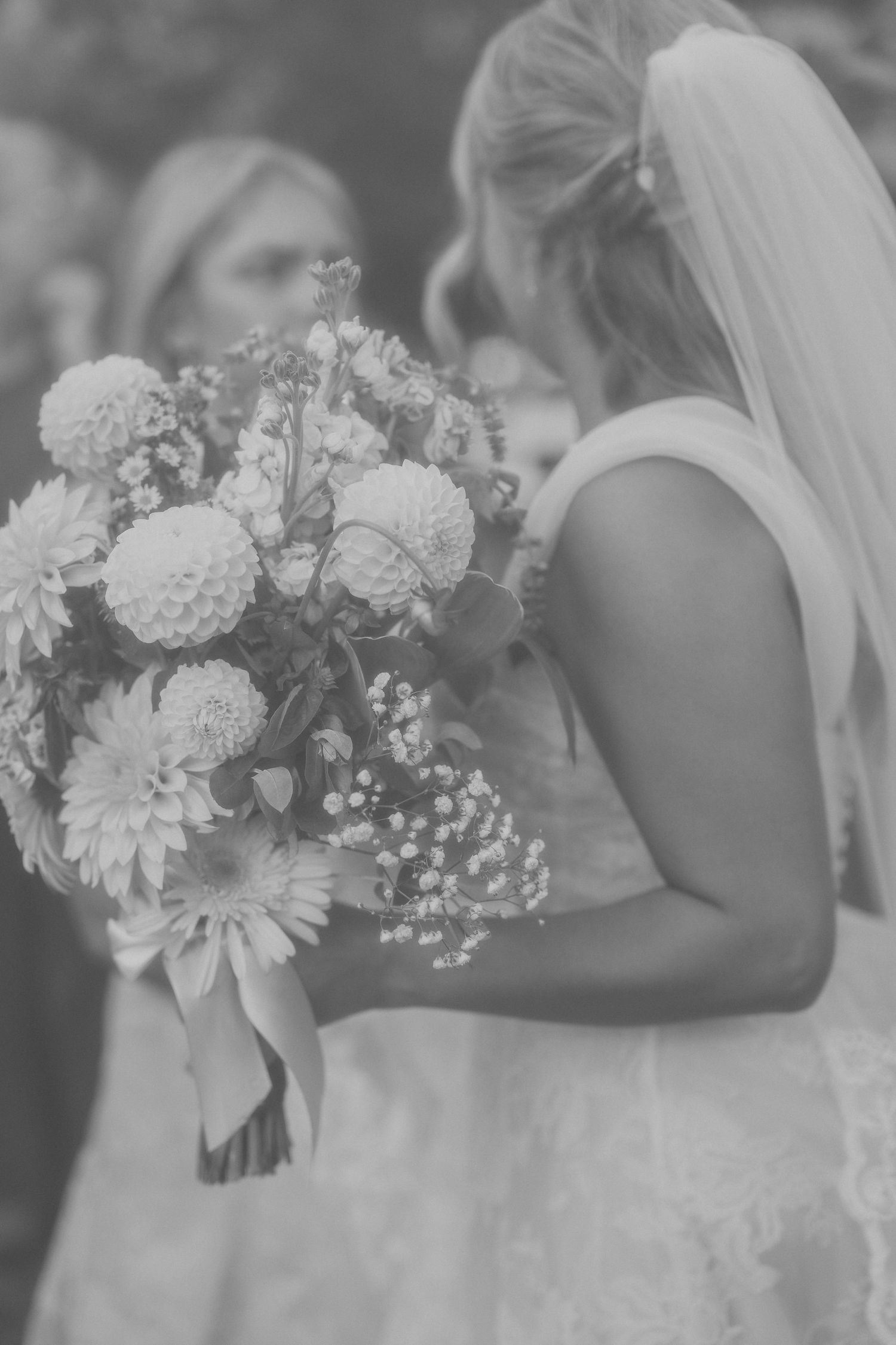 Black and white photo of a bride in a veil and lace dress holding a dahlia and baby's breath bouquet tied with ribbon while guests look on.