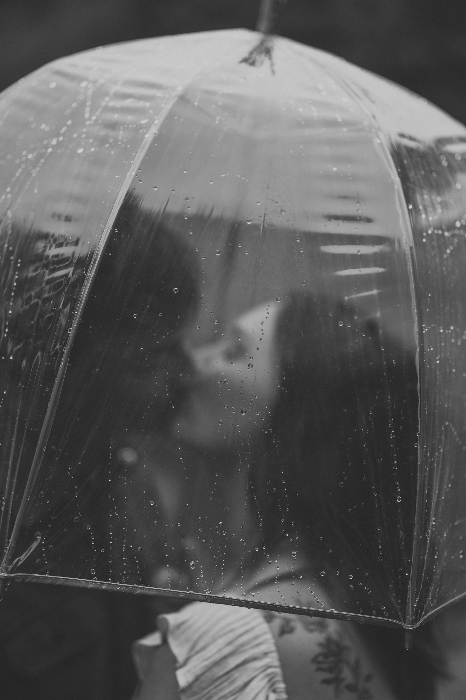 Black and white photo of a couple kissing behind a clear rain-speckled umbrella.