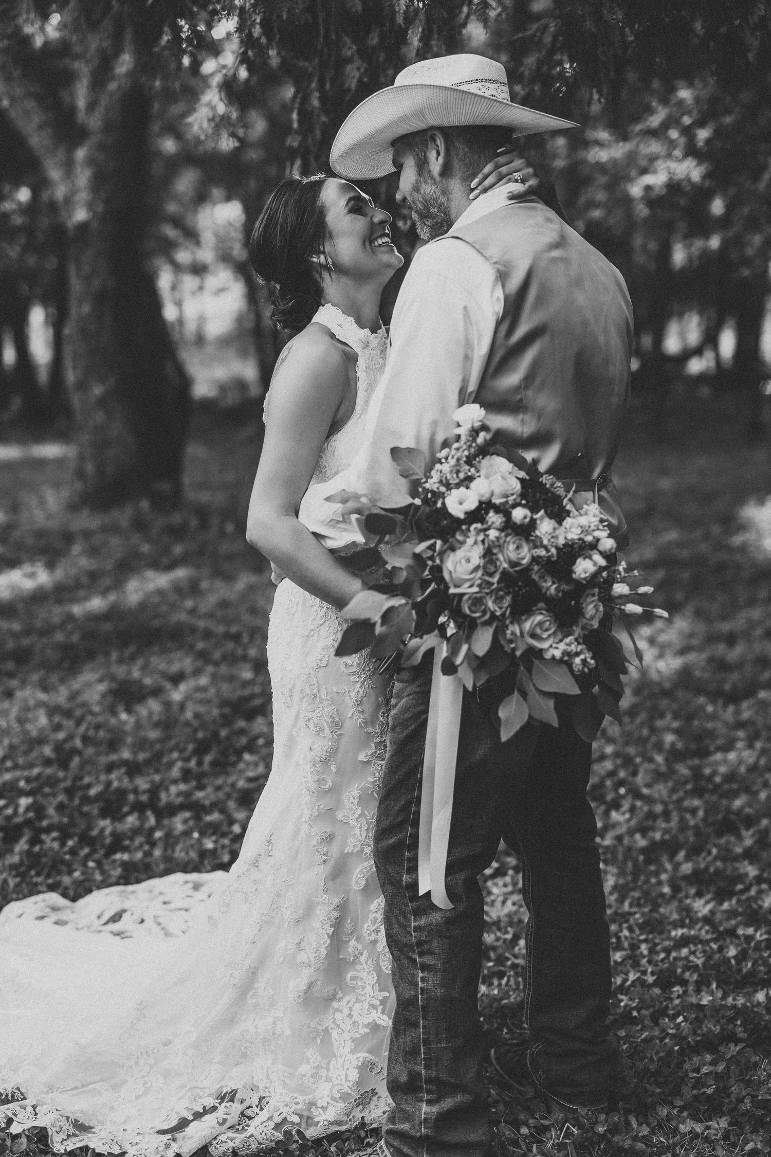 A couple in wedding attire embraces while holding a floral bouquet in a wooded setting.