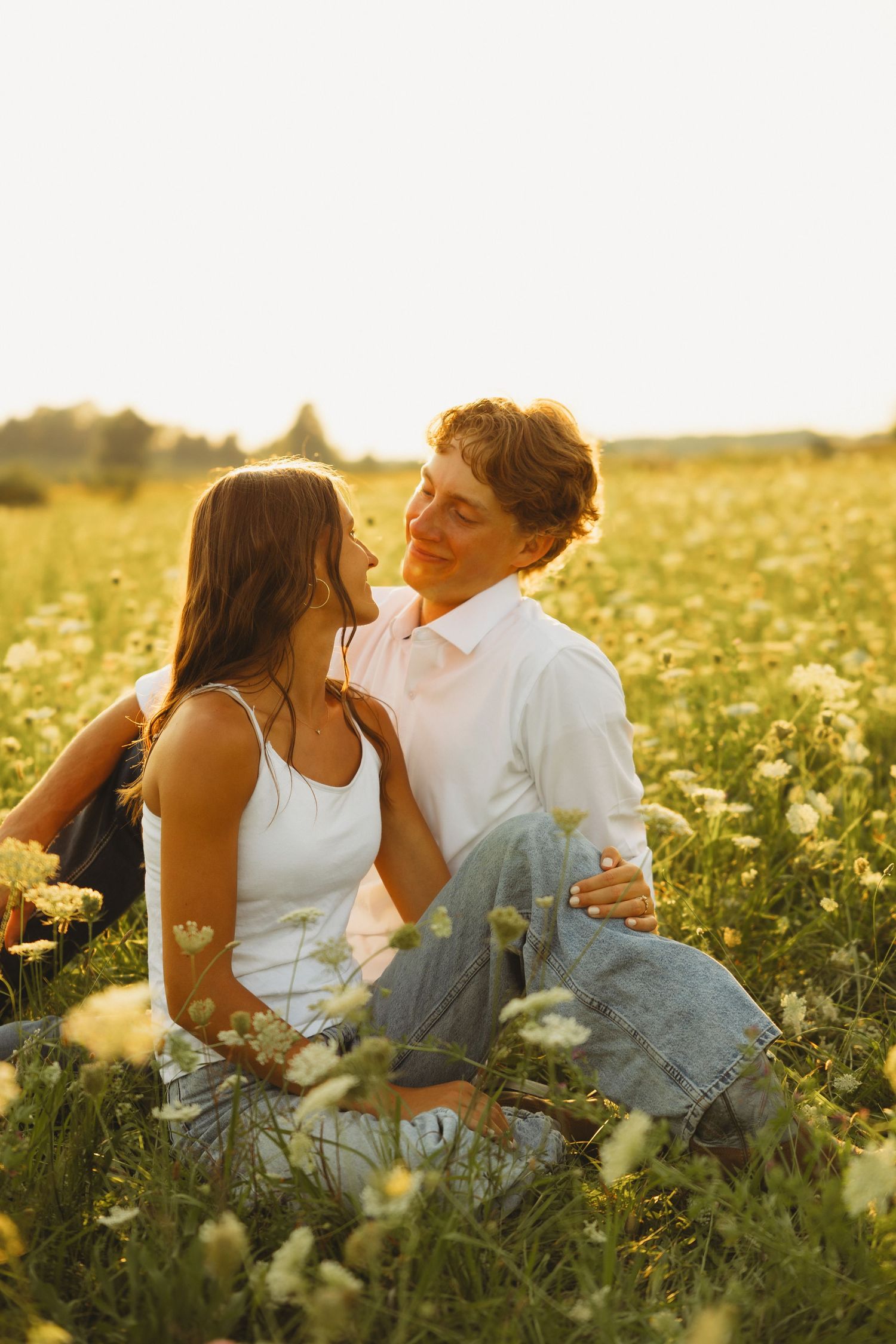Couple sitting in a sunny wildflower field, gazing into each other's eyes during golden hour.