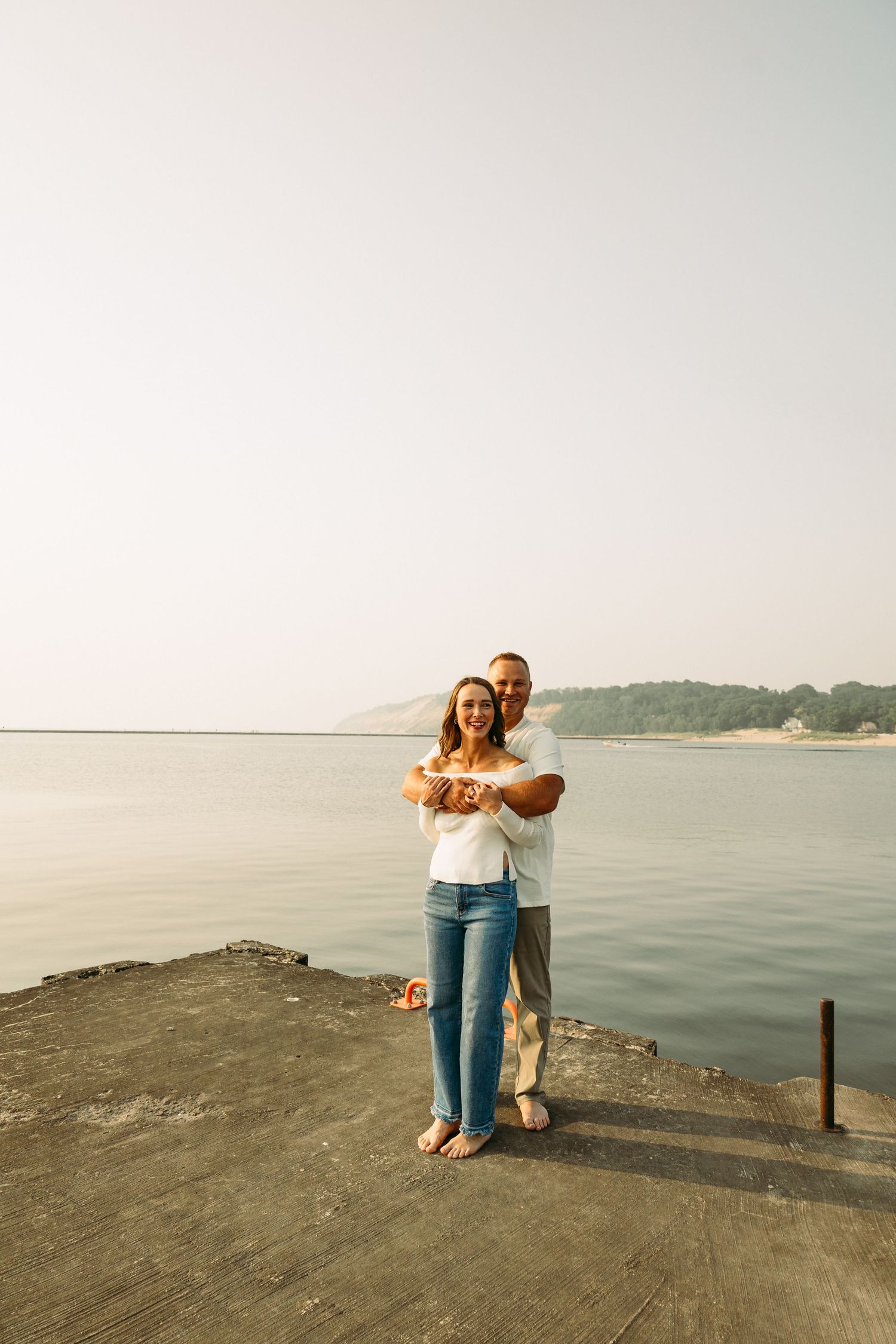 A couple embracing on a waterfront dock with calm water and hilly shoreline in the background.