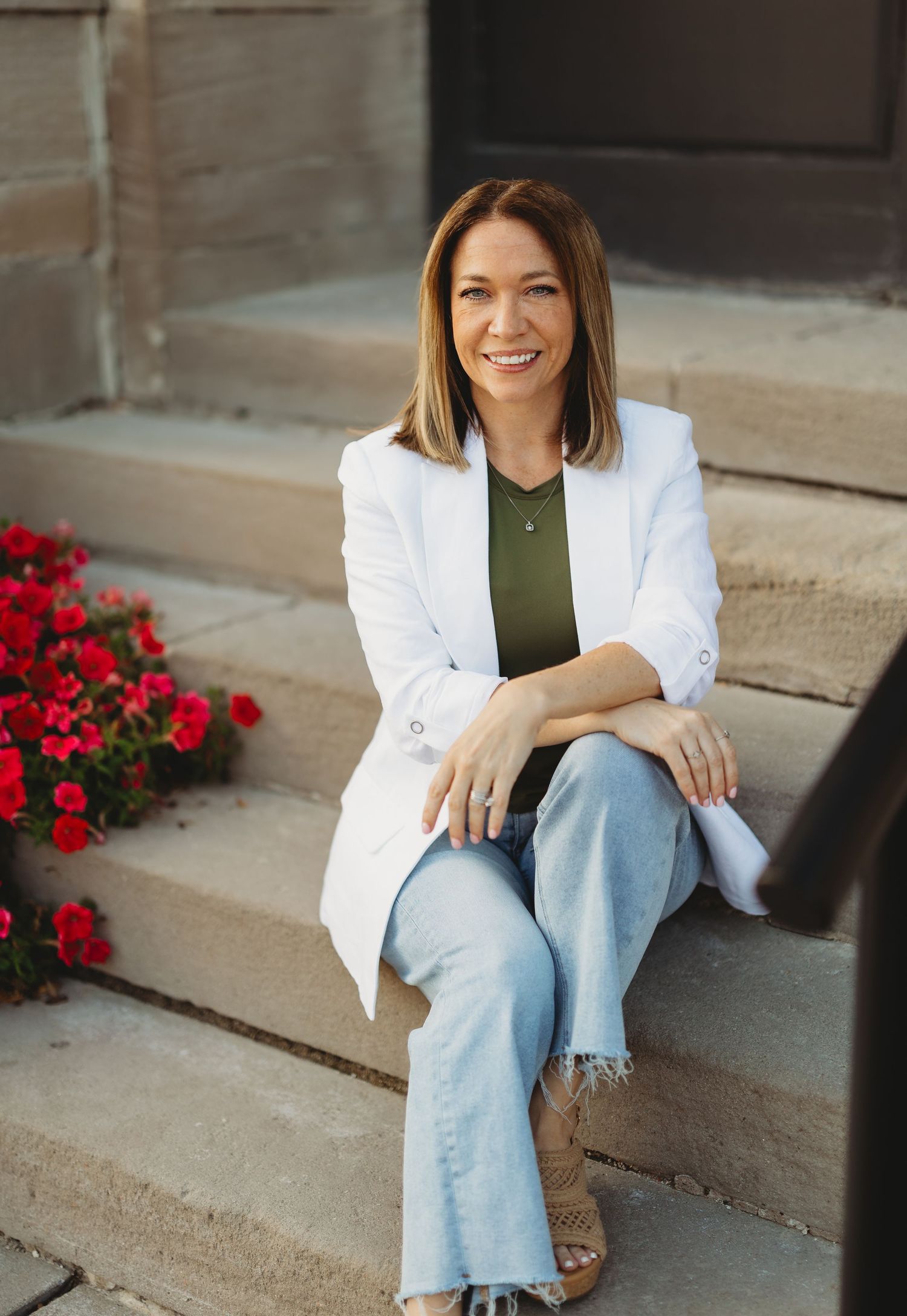 Smiling woman in white blazer, olive top, and jeans sitting on stone steps with red flowers nearby.