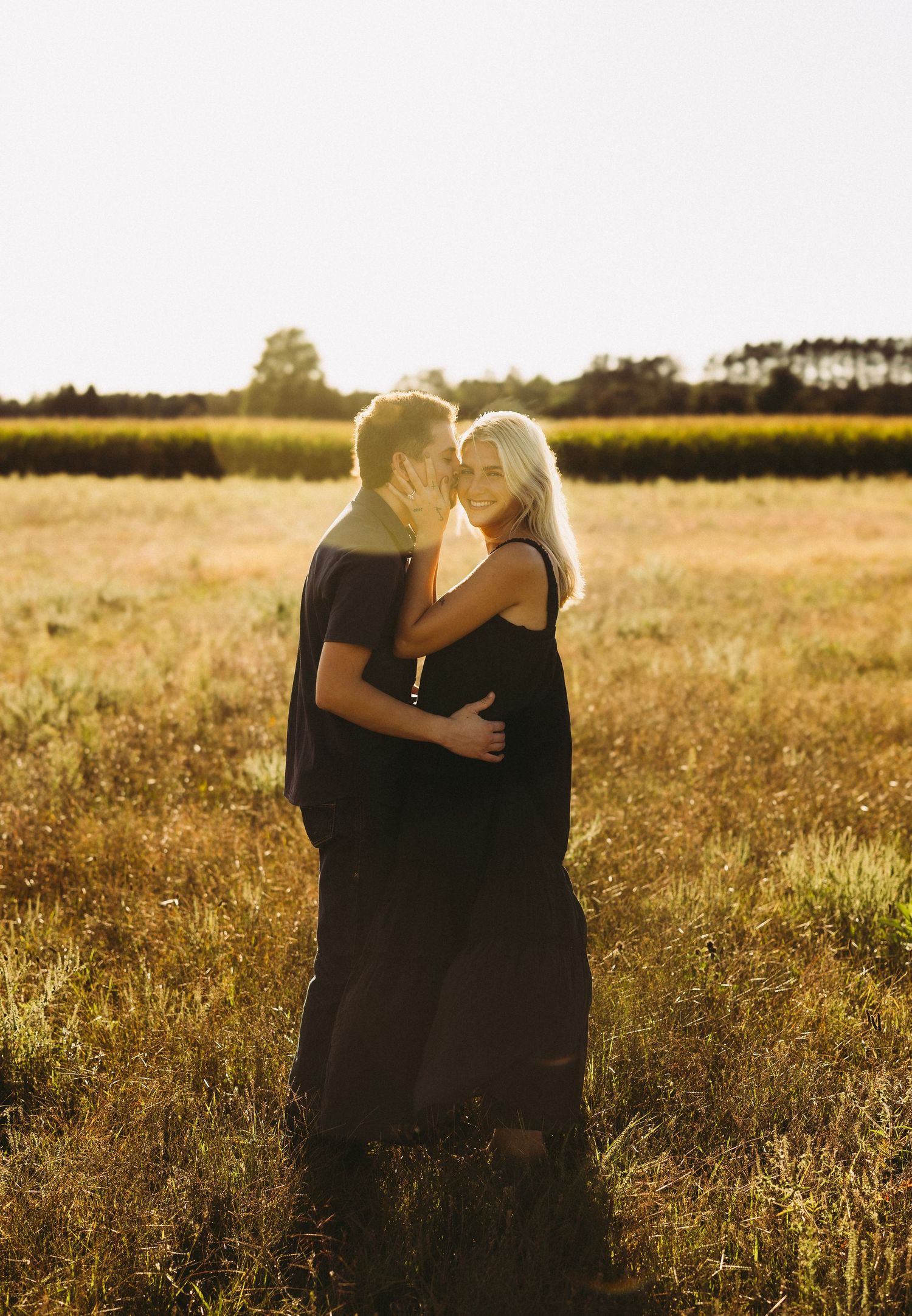 Couple embracing and kissing in a golden grass field at sunset, the woman in a black dress and the man in a dark shirt, with trees behind.