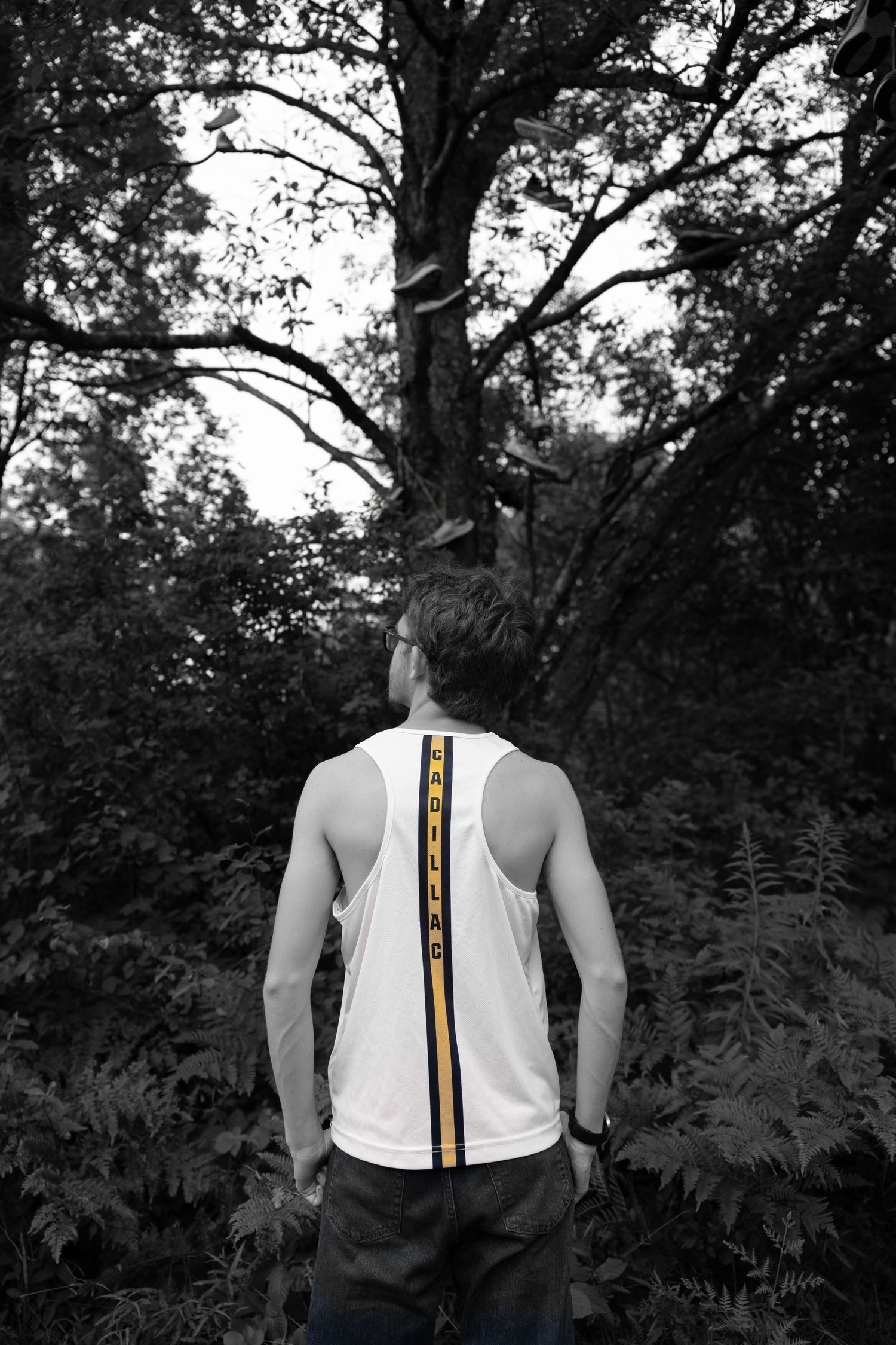 Black and white photo of a man in a white tank top with a yellow stripe, seen from behind, looking up at tall trees in a forest.