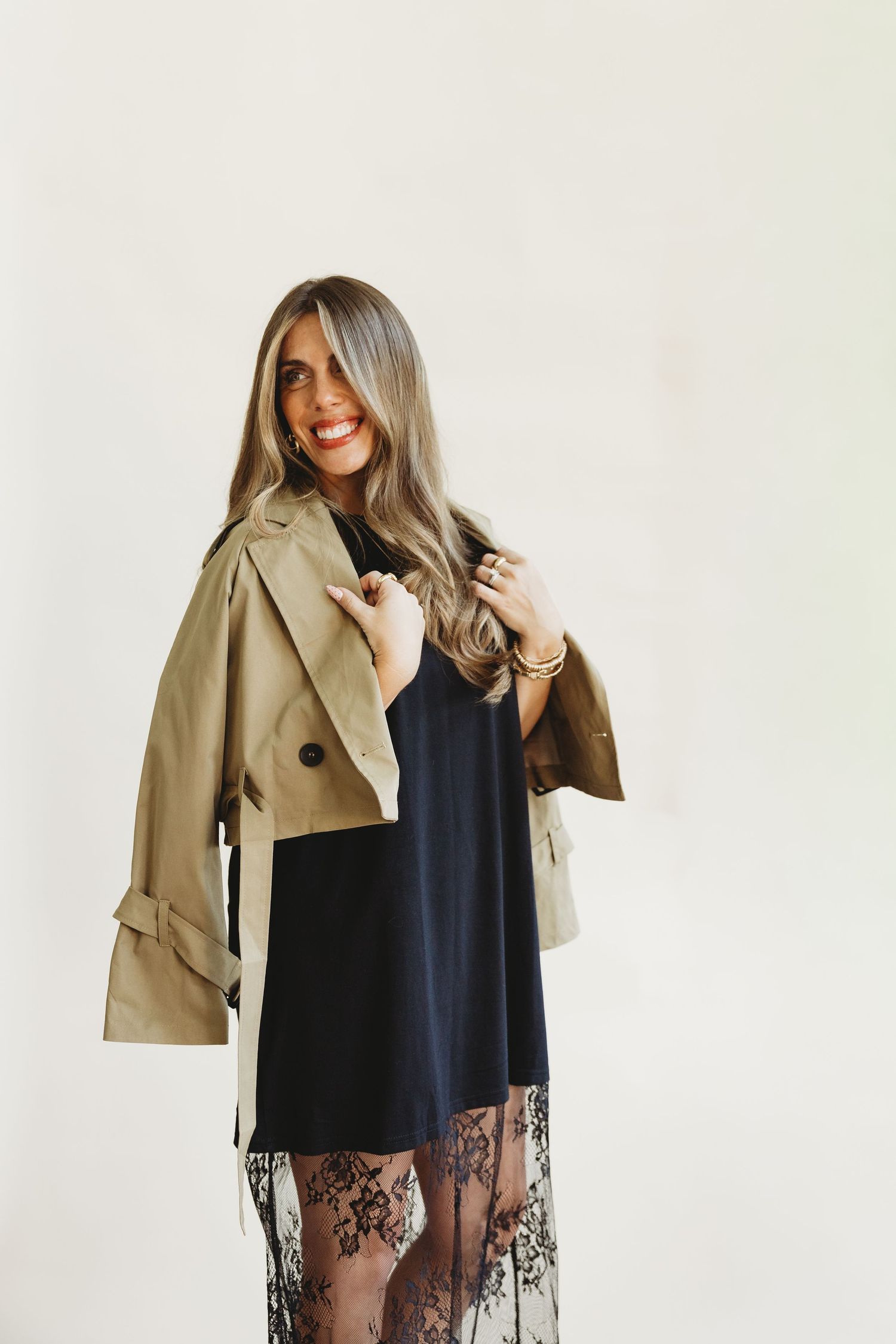 Smiling woman with long highlighted hair wears a cropped khaki trench jacket over her shoulders, a navy dress and a black lace skirt.