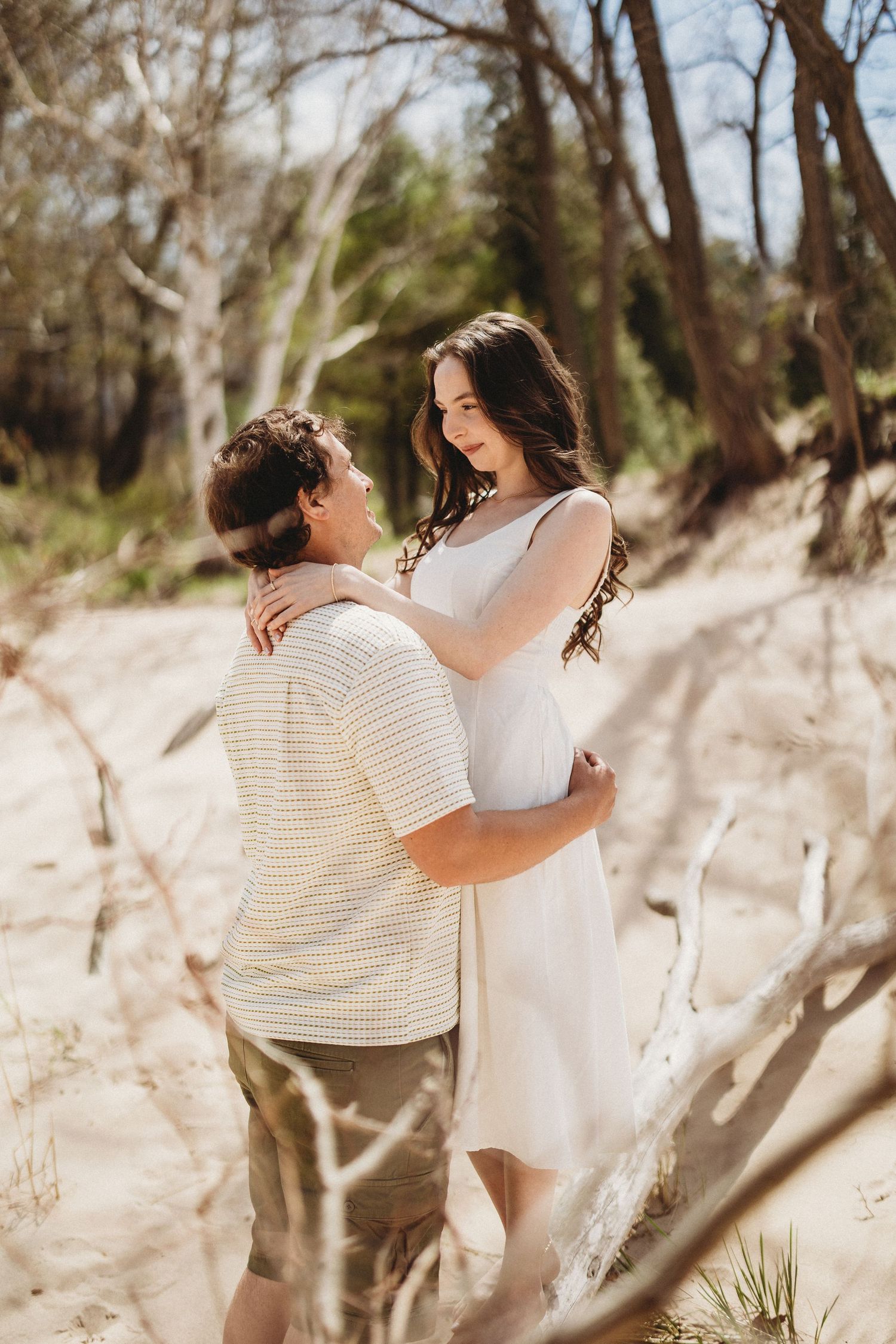 A couple embraces on a sandy beach trail surrounded by trees on a sunny afternoon.