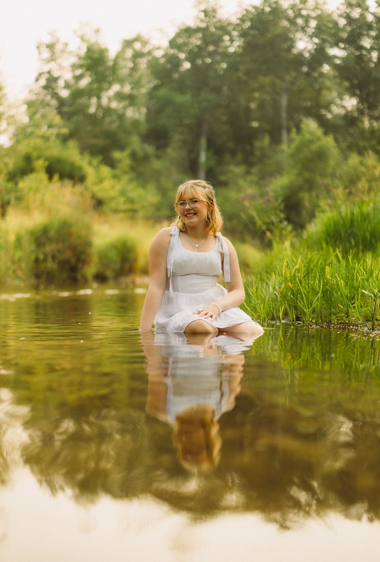 Figure in white dress sitting in reflective water surrounded by green vegetation.