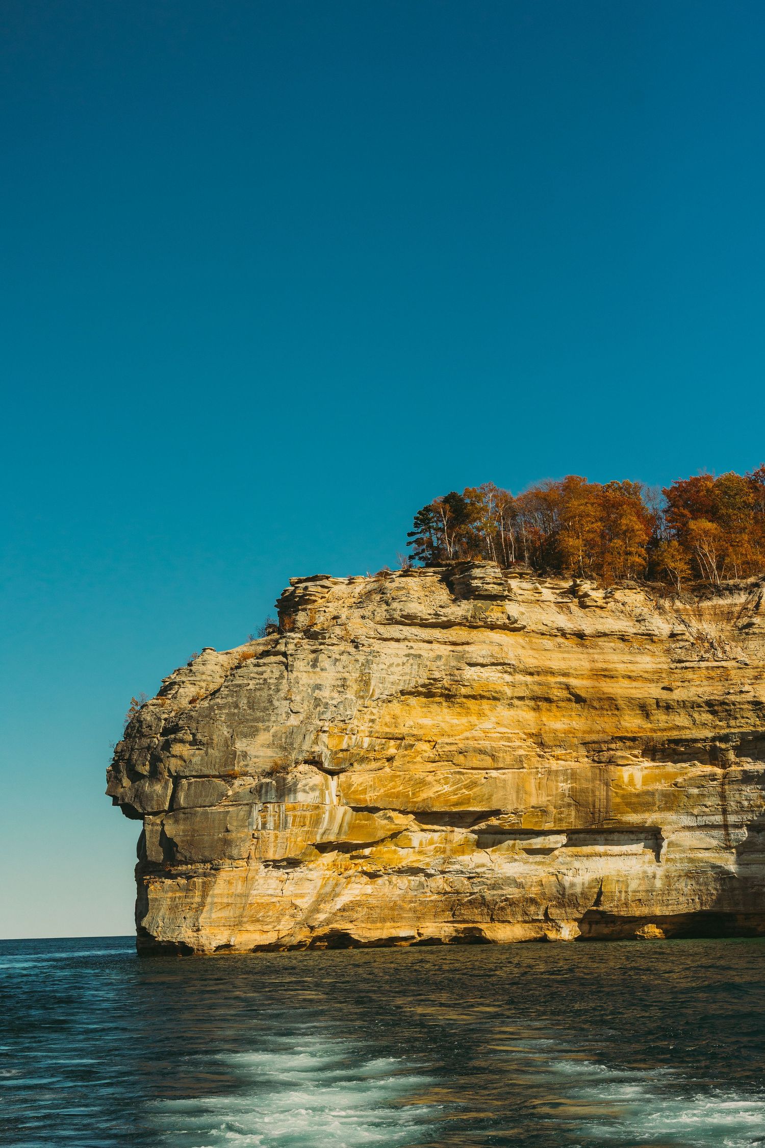 Layered sandstone cliff face with autumn trees on top against deep blue sky over Lake Superior.