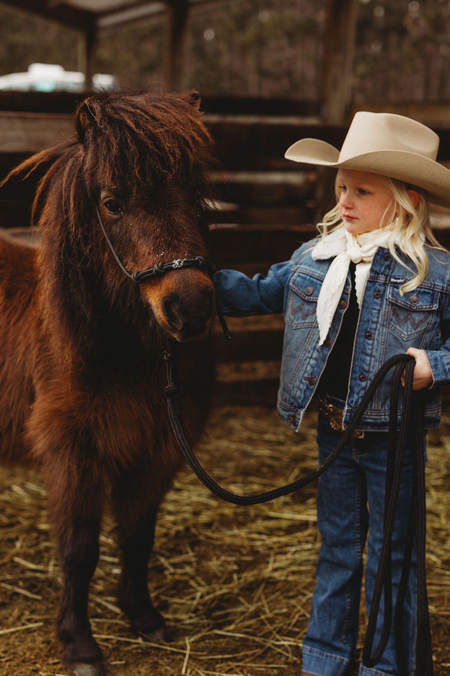 A person in western attire and denim jacket stands beside a brown pony in a straw-covered stable.