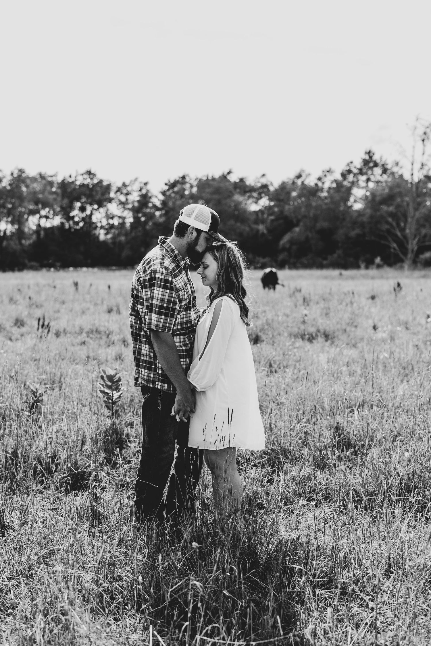 A couple embraces in a serene black and white meadow scene with tall grass and trees in the background.