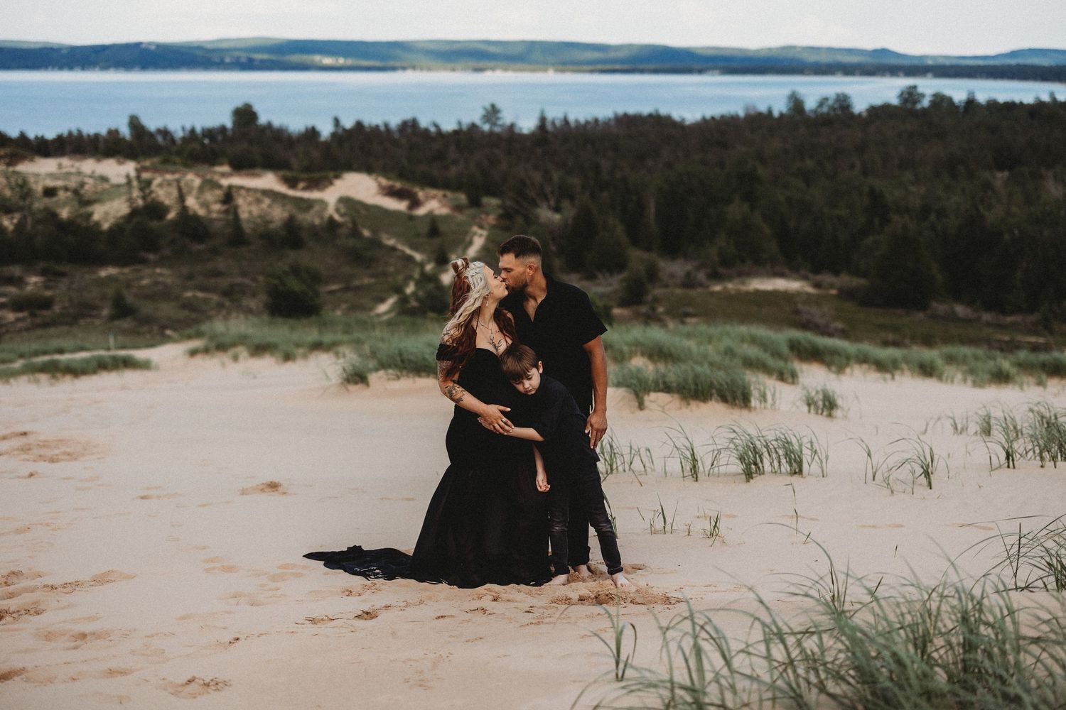 A couple in black outfits embraces on sandy dunes overlooking a lake and forest landscape at dusk.