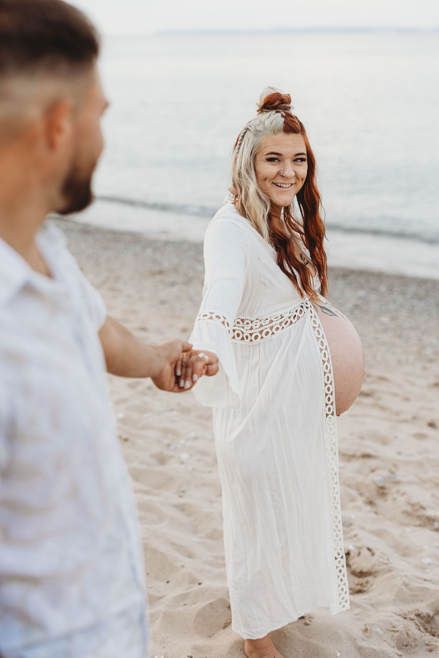 Person in white lace dress twirling on beach during engagement photoshoot.
