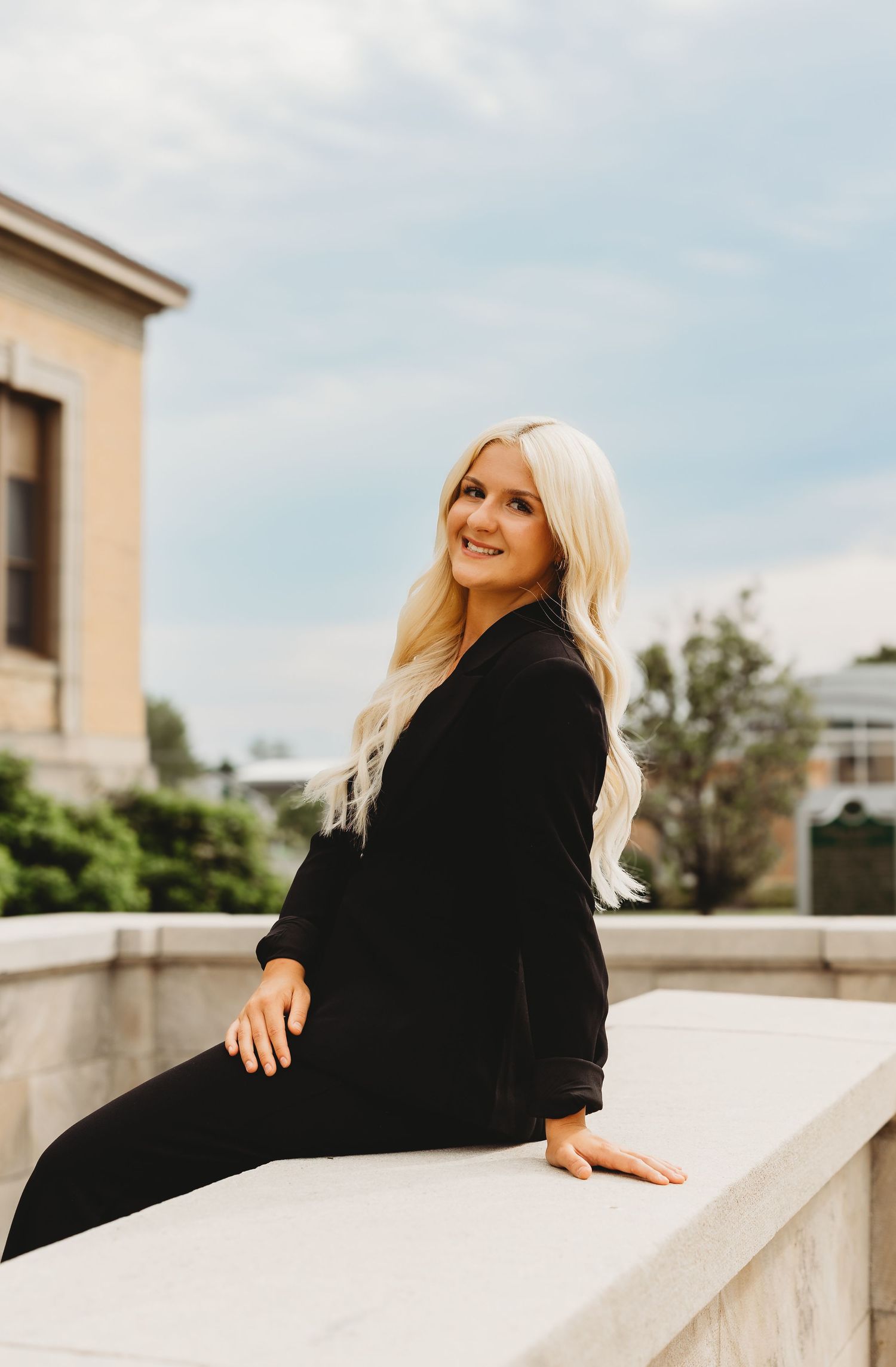 Smiling blonde woman in black outfit sitting on stone ledge outdoors with building and cloudy sky background.