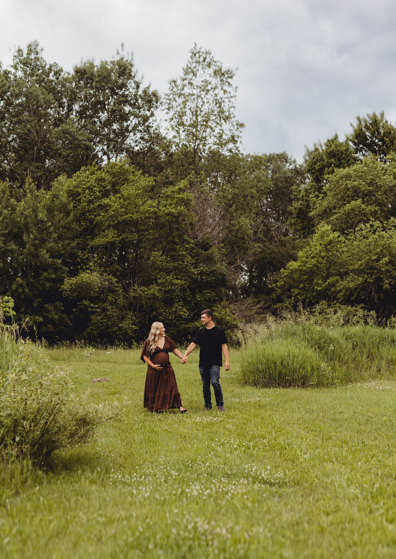 Pregnant couple holding hands in a lush green meadow, woman in burgundy dress, man in black shirt and jeans.
