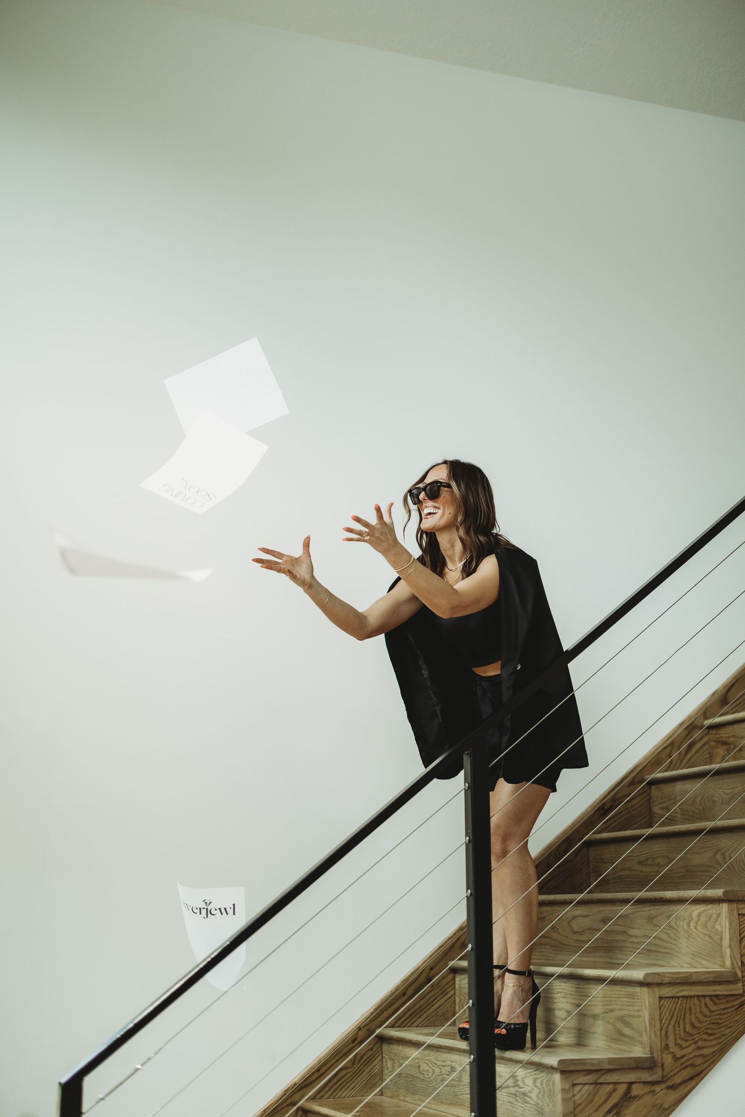 Woman in black outfit tossing papers in the air on modern staircase, wearing sunglasses, celebratory pose.