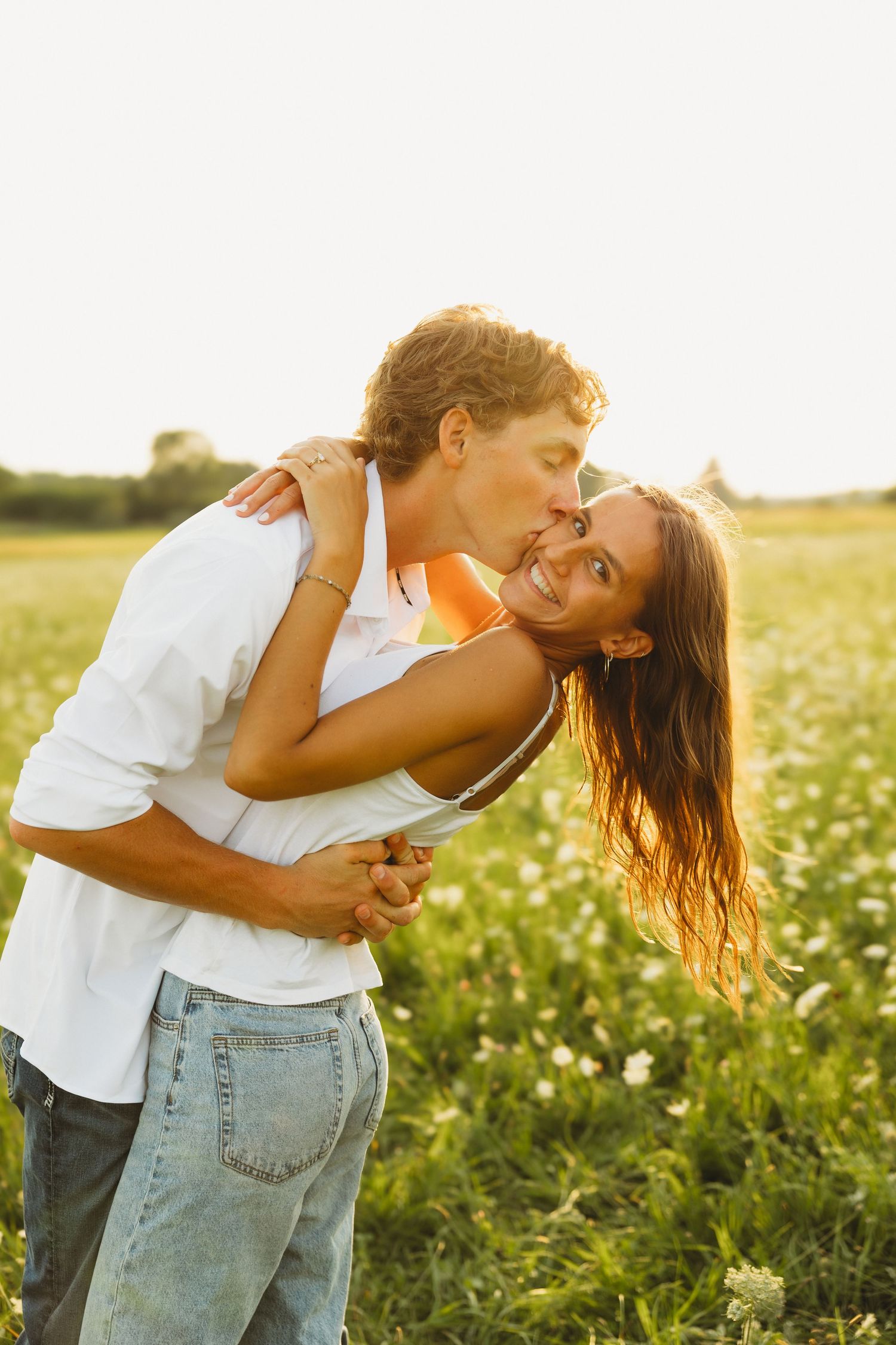 Couple sharing a romantic kiss in a sunny wildflower field, man in white shirt and jeans embracing smiling woman.